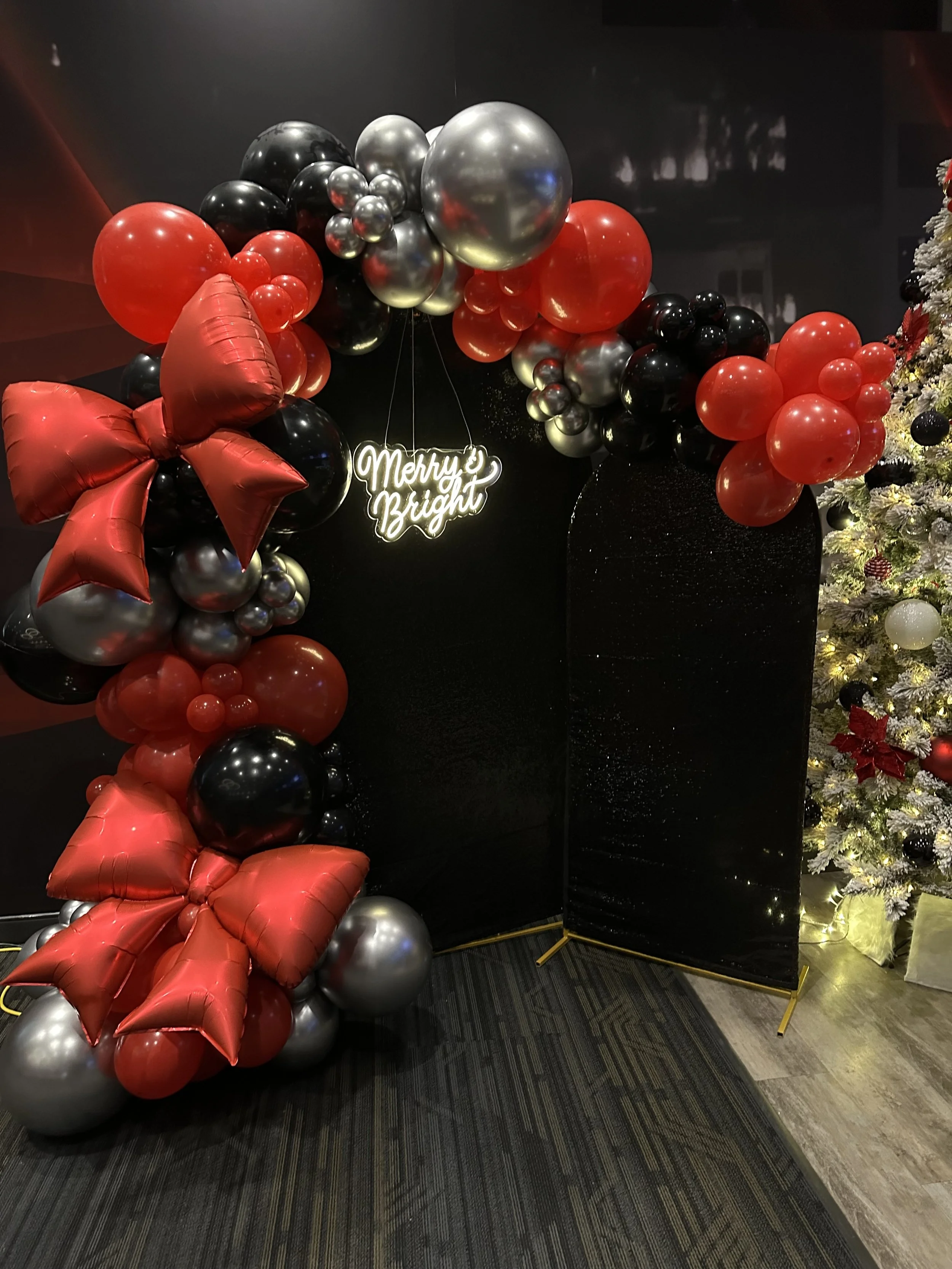 Decorative holiday scene with red, black, and silver balloons forming an arch around a black backdrop, with a neon sign saying 'Merry & Bright'. A Christmas tree with ornaments and lights is visible on the right.