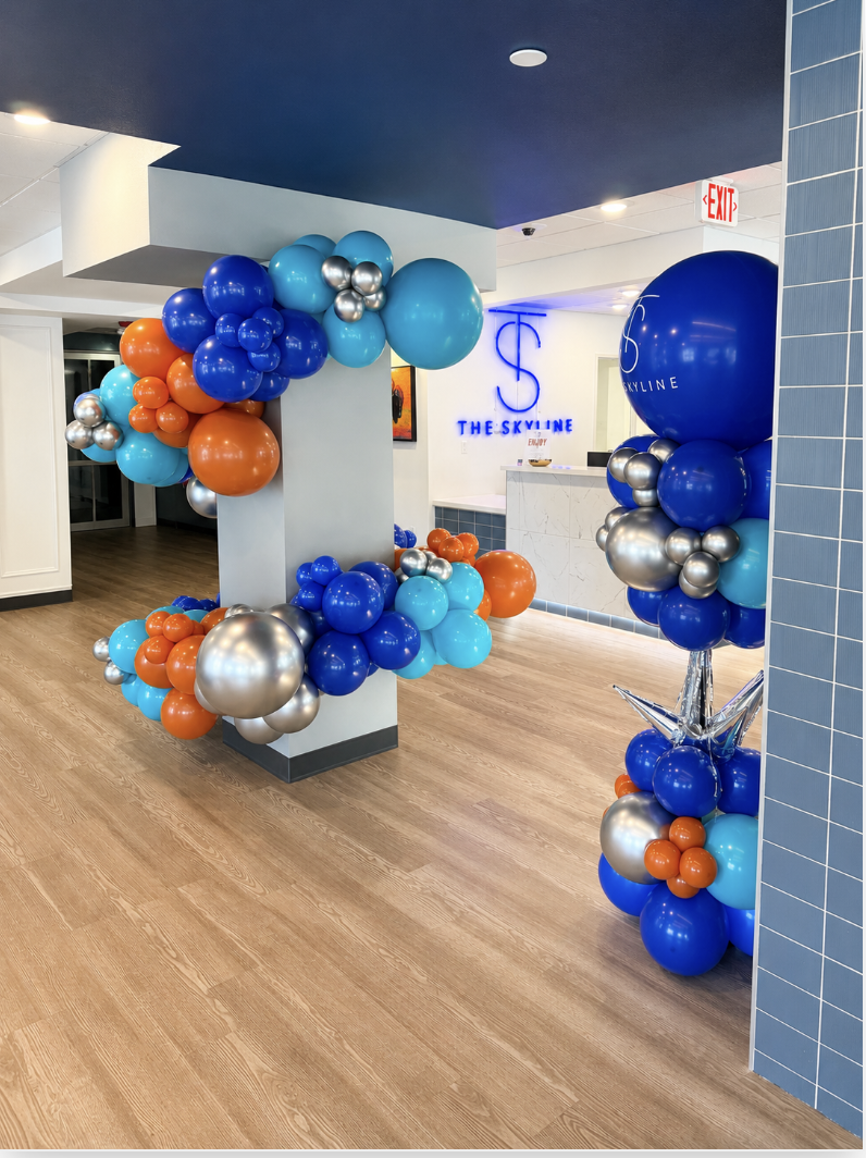 Decorative balloon arrangements in blue, orange, and silver at the entrance of a modern office or venue, with a sign reading "The Skyline" in blue neon lights on the wall.