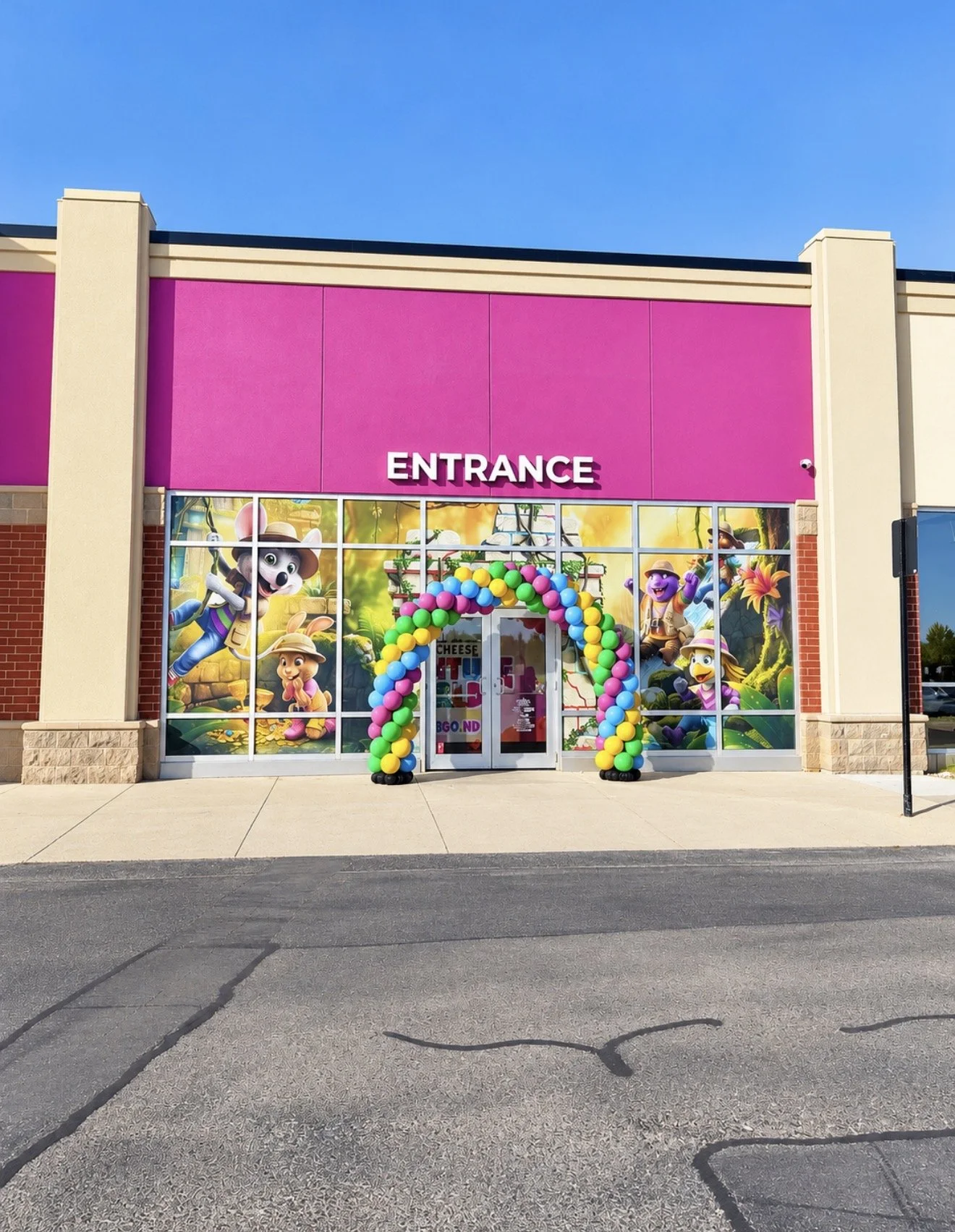 Colorful storefront with pink and cream exterior, large windows with cartoon animal characters and tropical jungle background, balloon arch at entrance, and the word "ENTRANCE" sign above.