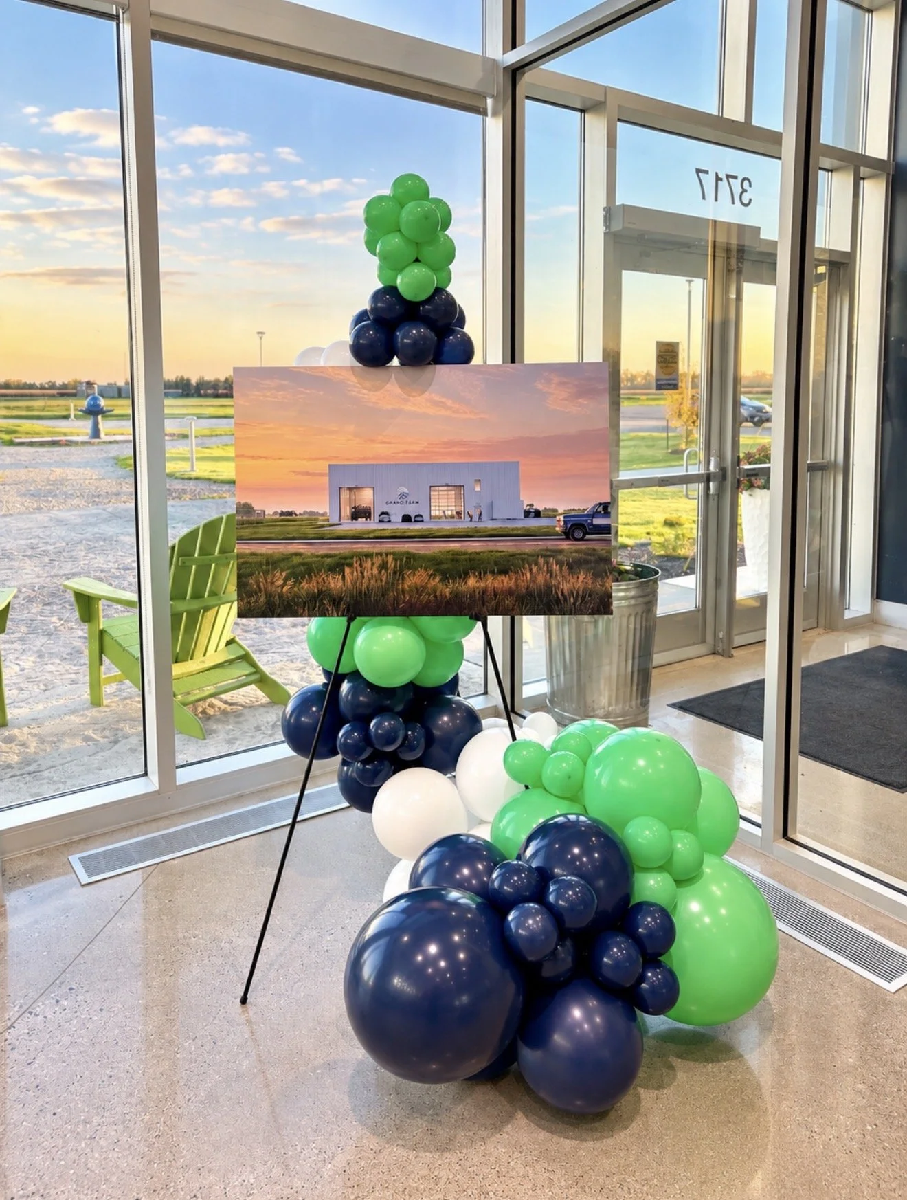 Indoor display of green, navy blue, and white balloons arranged around a large photo of a modern building with a sunset sky, inside a building with large glass windows, a green outdoor chair visible, and a view of a parking lot and grassy landscape o