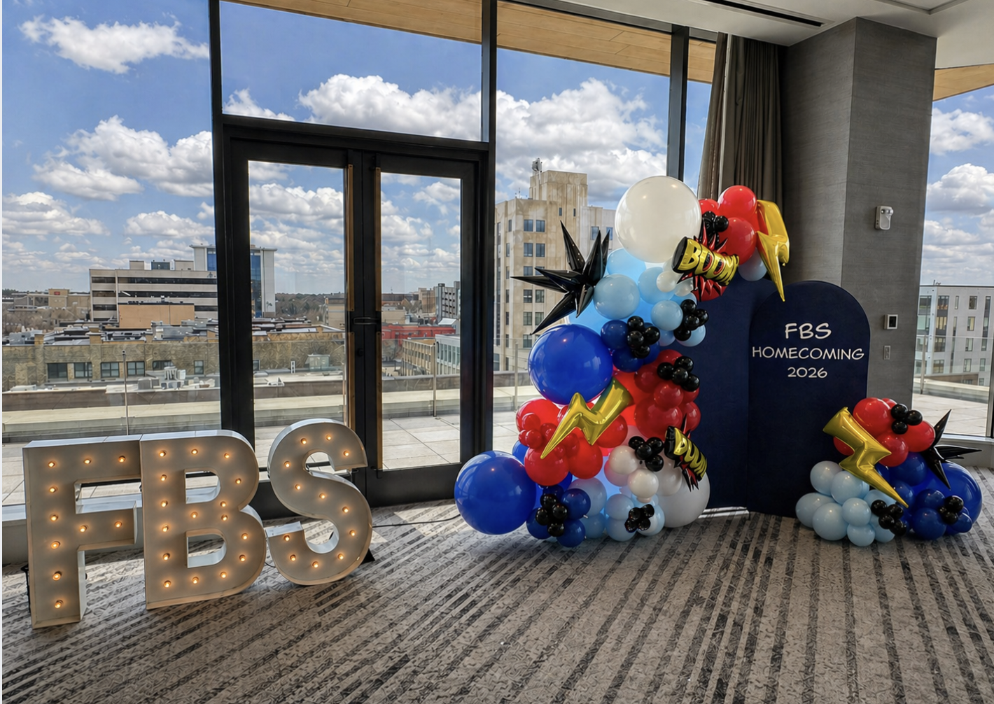 Balloon display for FBS homecoming 2026 with large illuminated 'FBS' letters in the foreground, and colorful balloons arranged on a backdrop with a city skyline and cloudy sky visible through large windows.