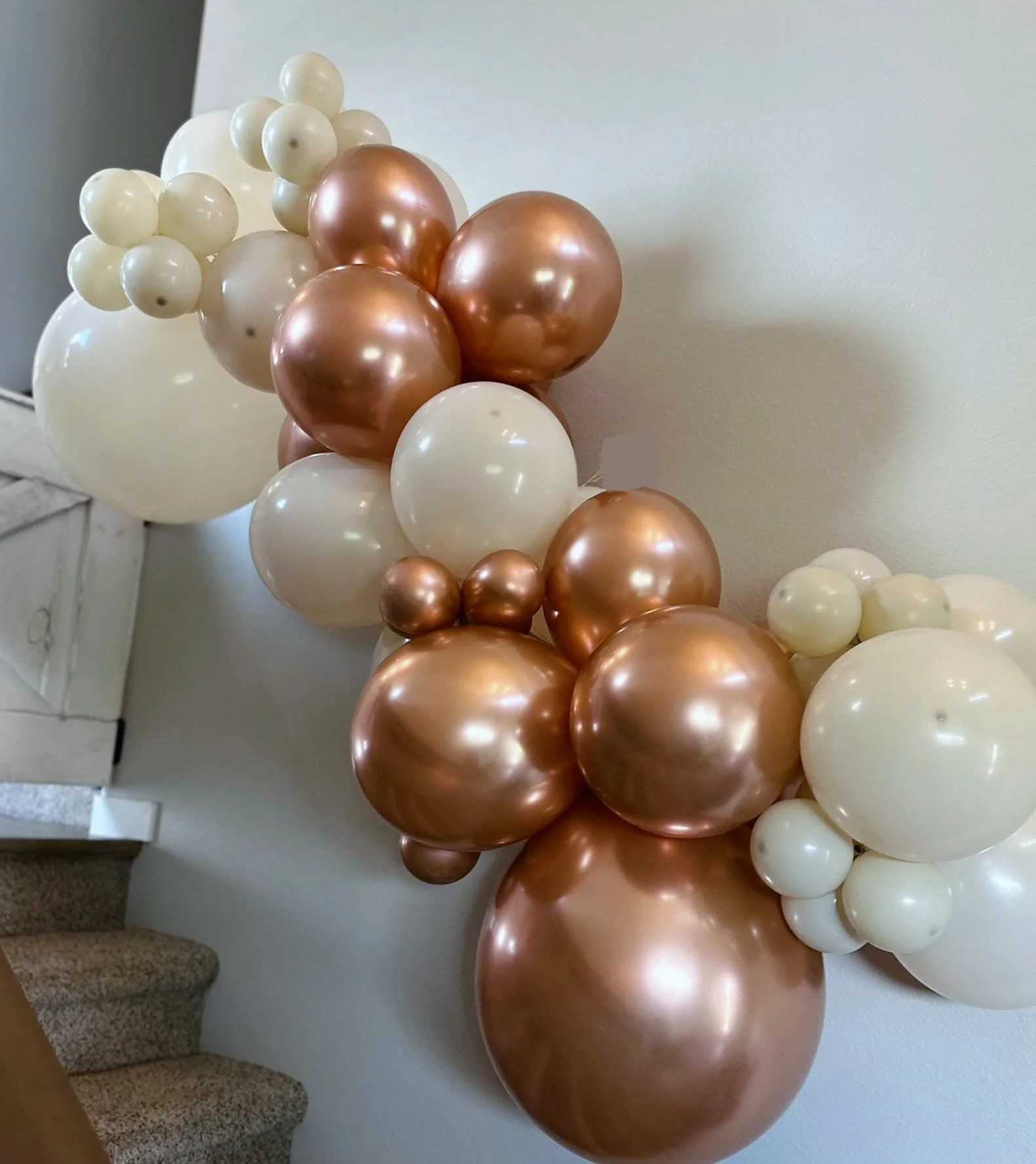 A decorative arrangement of white and metallic rose gold balloons near a staircase and a white wall.