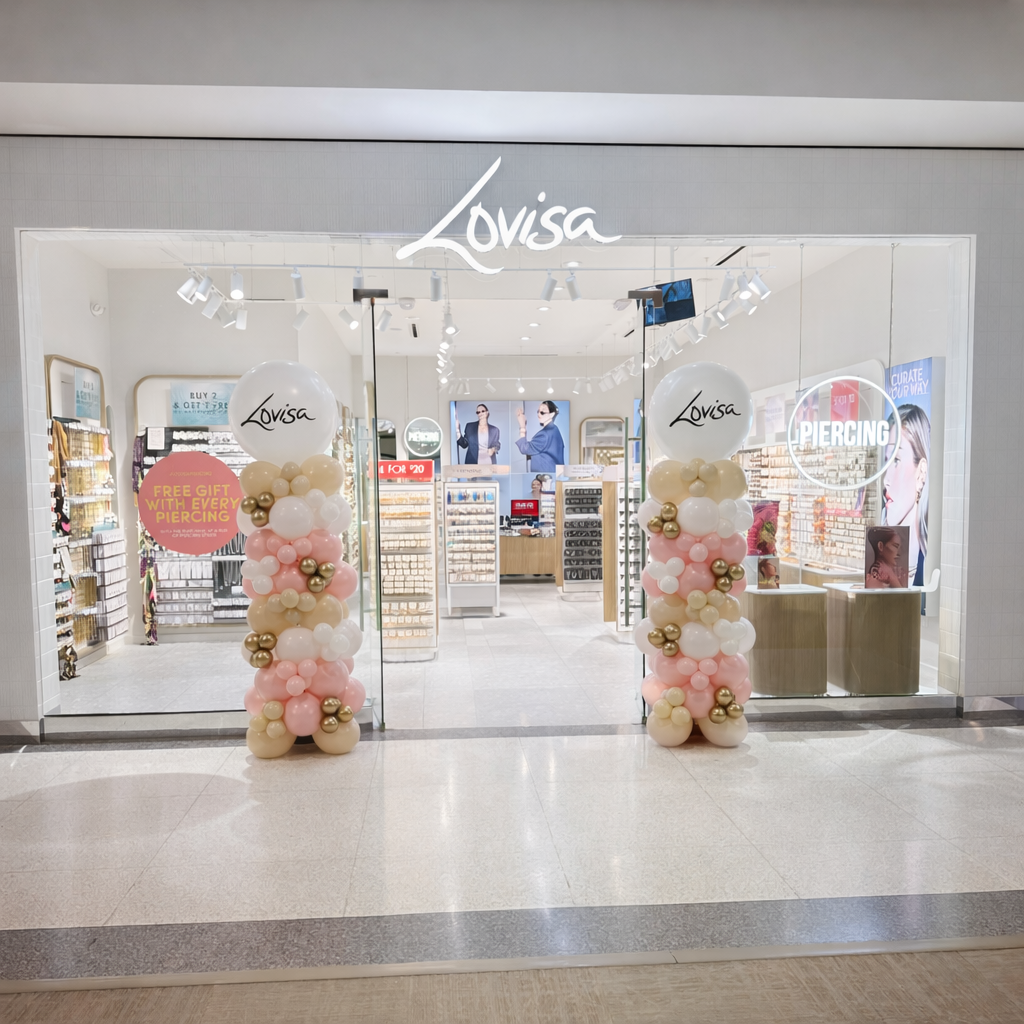Front view of a retail store named Louisa with balloon columns at entrance, promotional sign for free gift with piercing, and digital screens inside advertising jewelry and piercing services.