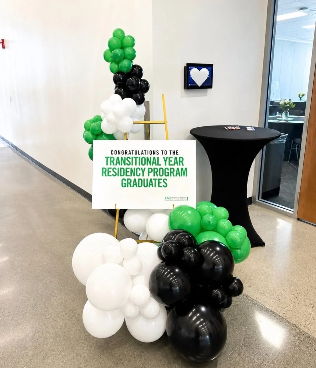Balloon display with white, black, and green clusters, a sign that reads 'Congratulations to the Transitional Year Residency Program Graduates,' a black high table, and a monitor with a white heart symbol on the wall in the background.