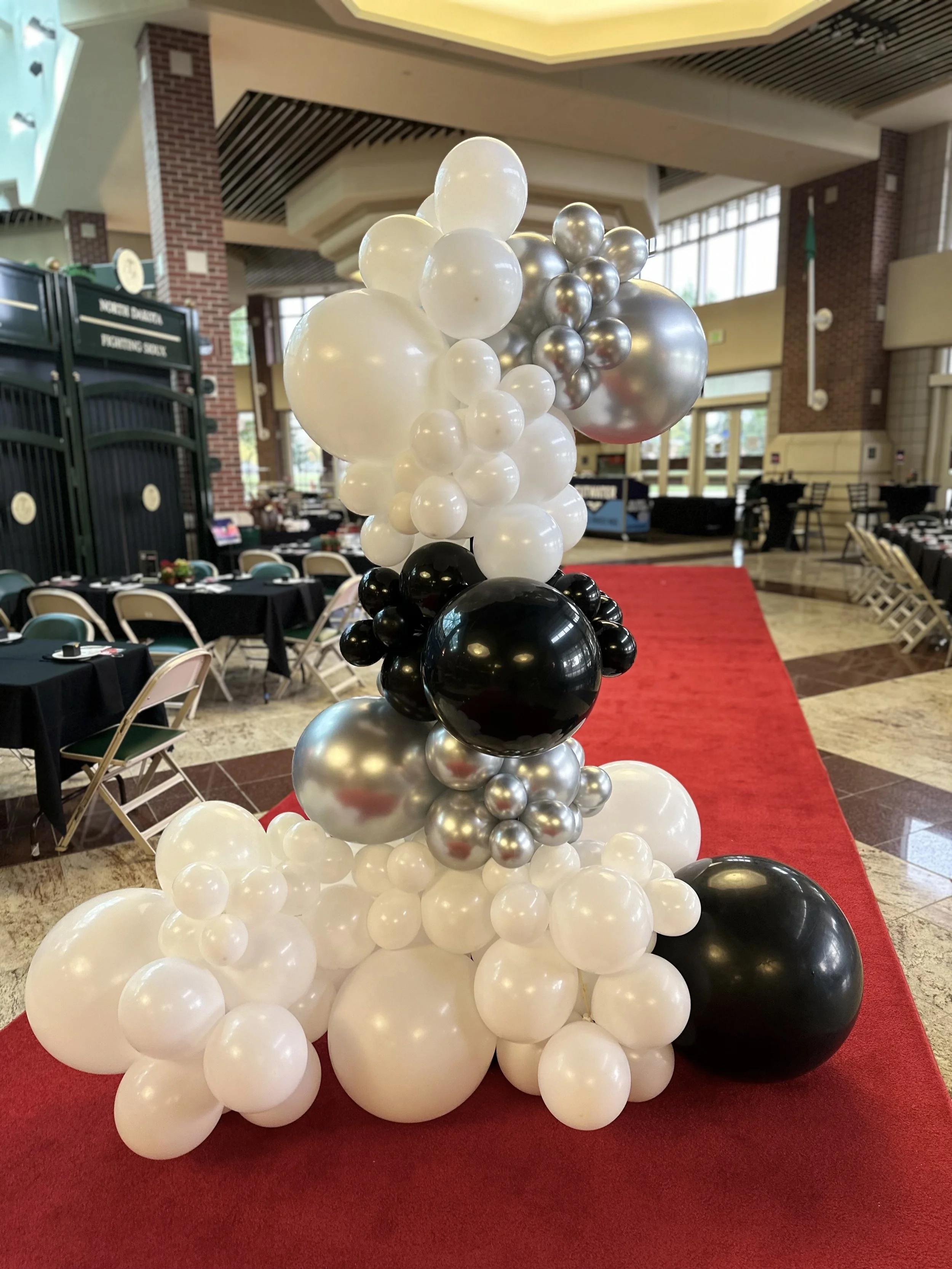A large balloon arrangement with black, white, silver, and grey balloons in various sizes, set on a red carpet in a decorated indoor event space with tables covered in black tablecloths.