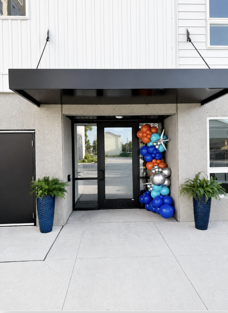 Decorative balloon arrangement with orange, blue, silver, and teal balloons flanking the glass entrance door of a modern building, with potted green plants in blue vases on either side.