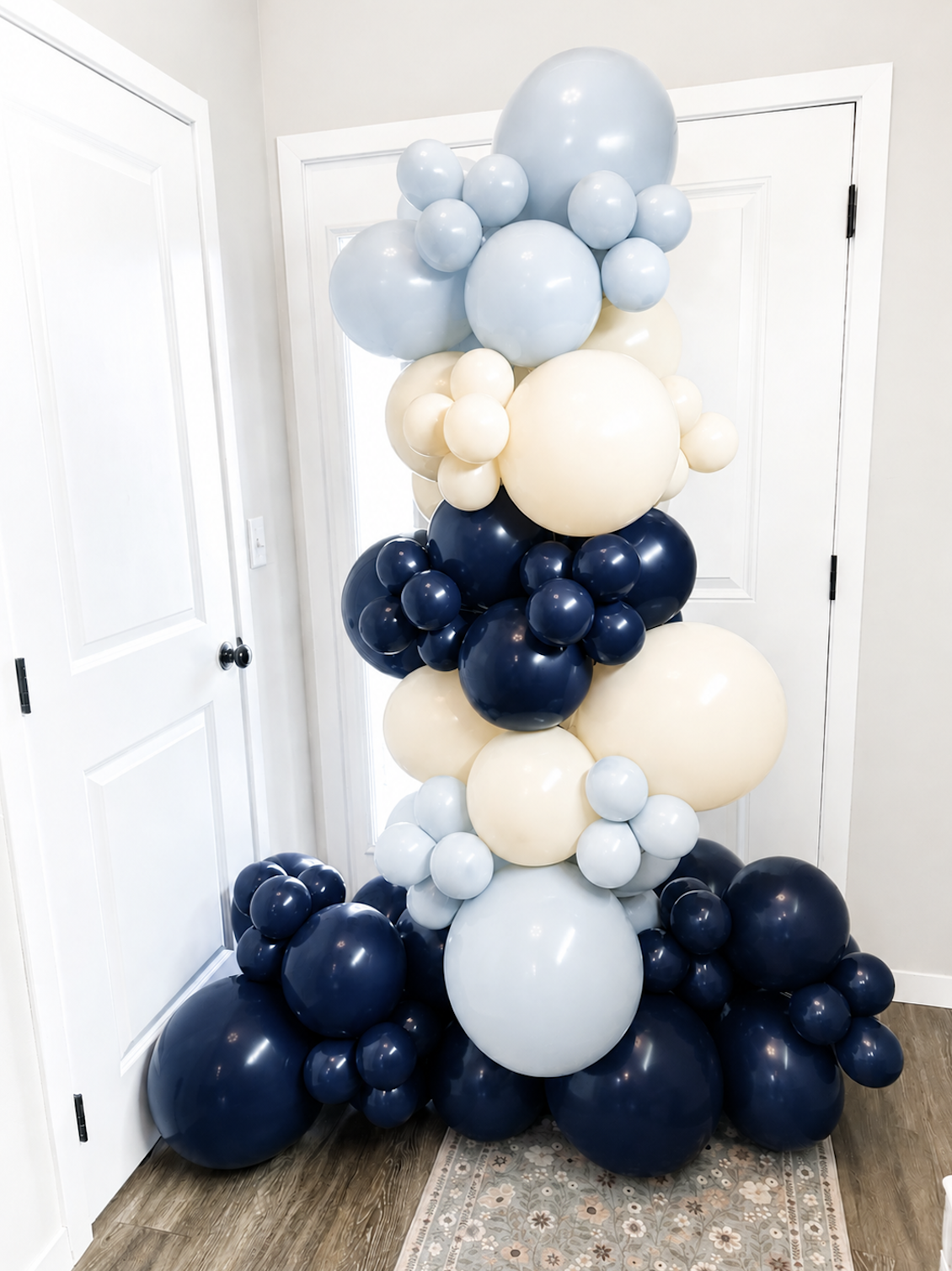 A large balloon arrangement with shades of blue, white, and cream balloons stacked and clustered beside white double doors in a room.