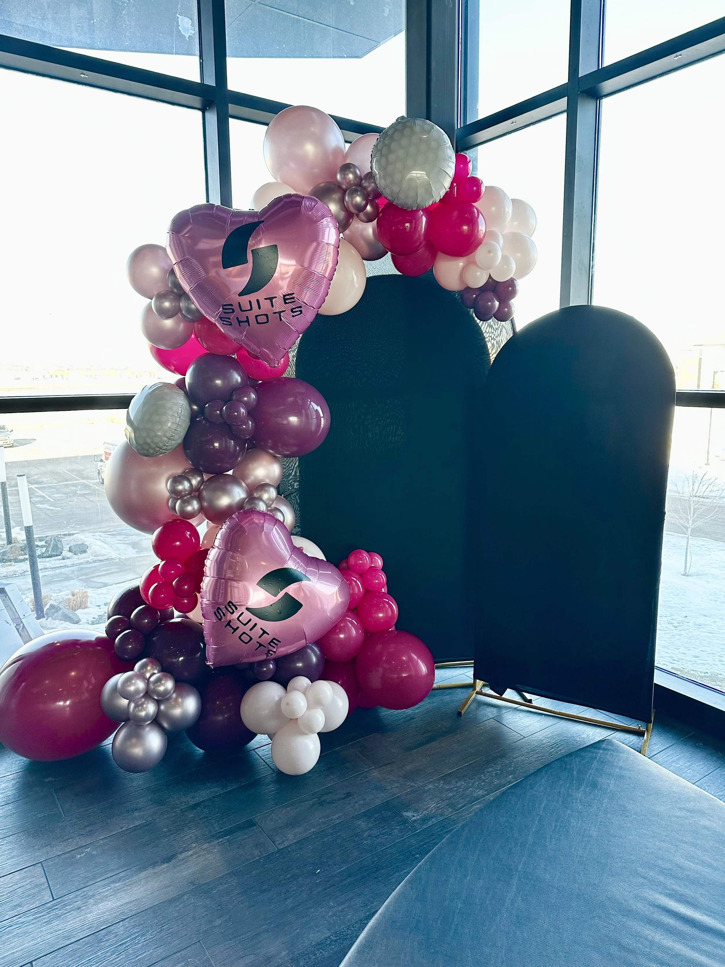 A decorative balloon arrangement with pink, white, purple, and metallic balloons next to a black photo booth with a large round top, placed in Suite Shots Fargo North Dakota
