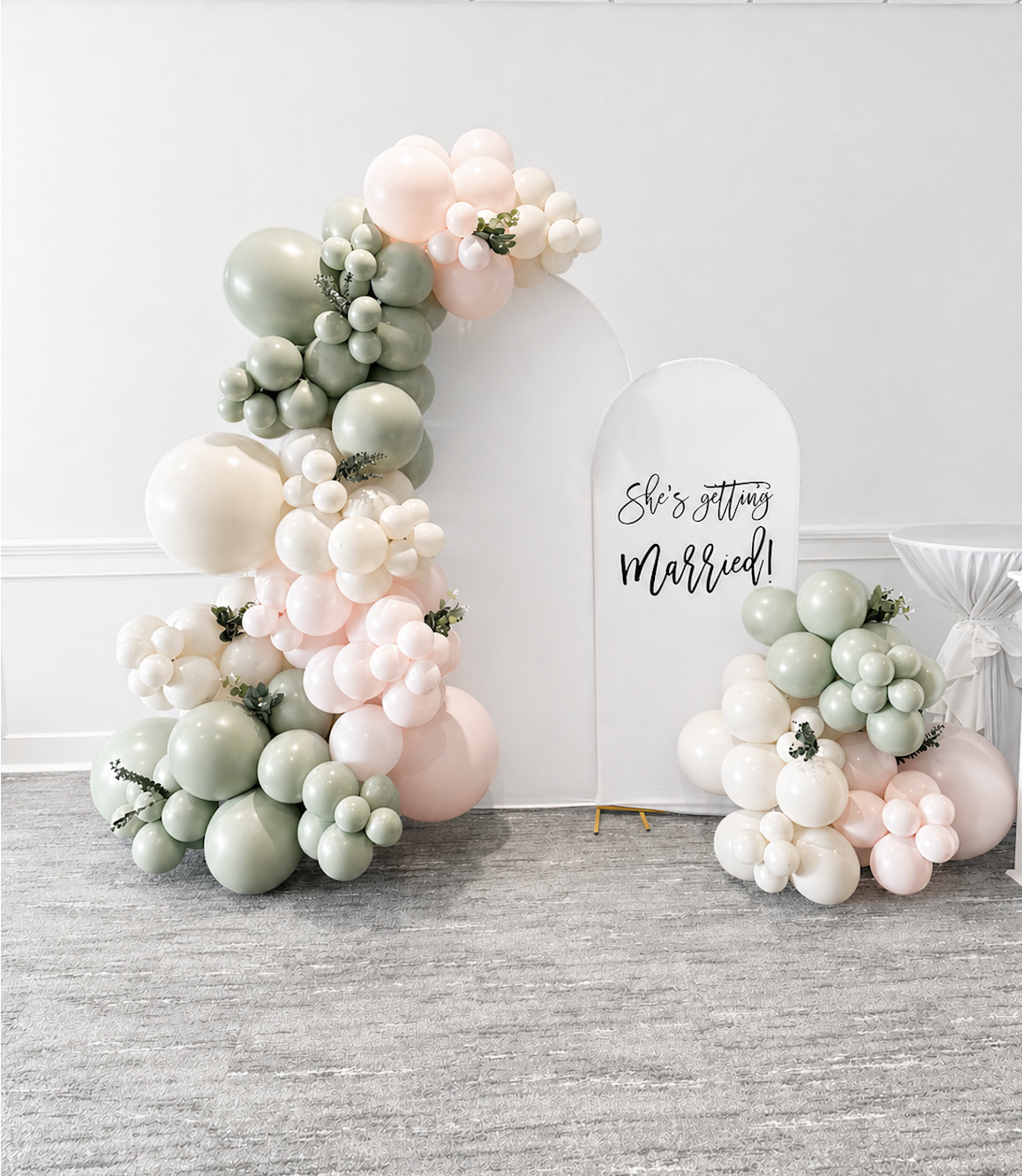 Balloon display with pastel colors and greenery, arching around a white sign that reads "She's getting married!" for a wedding celebration.
