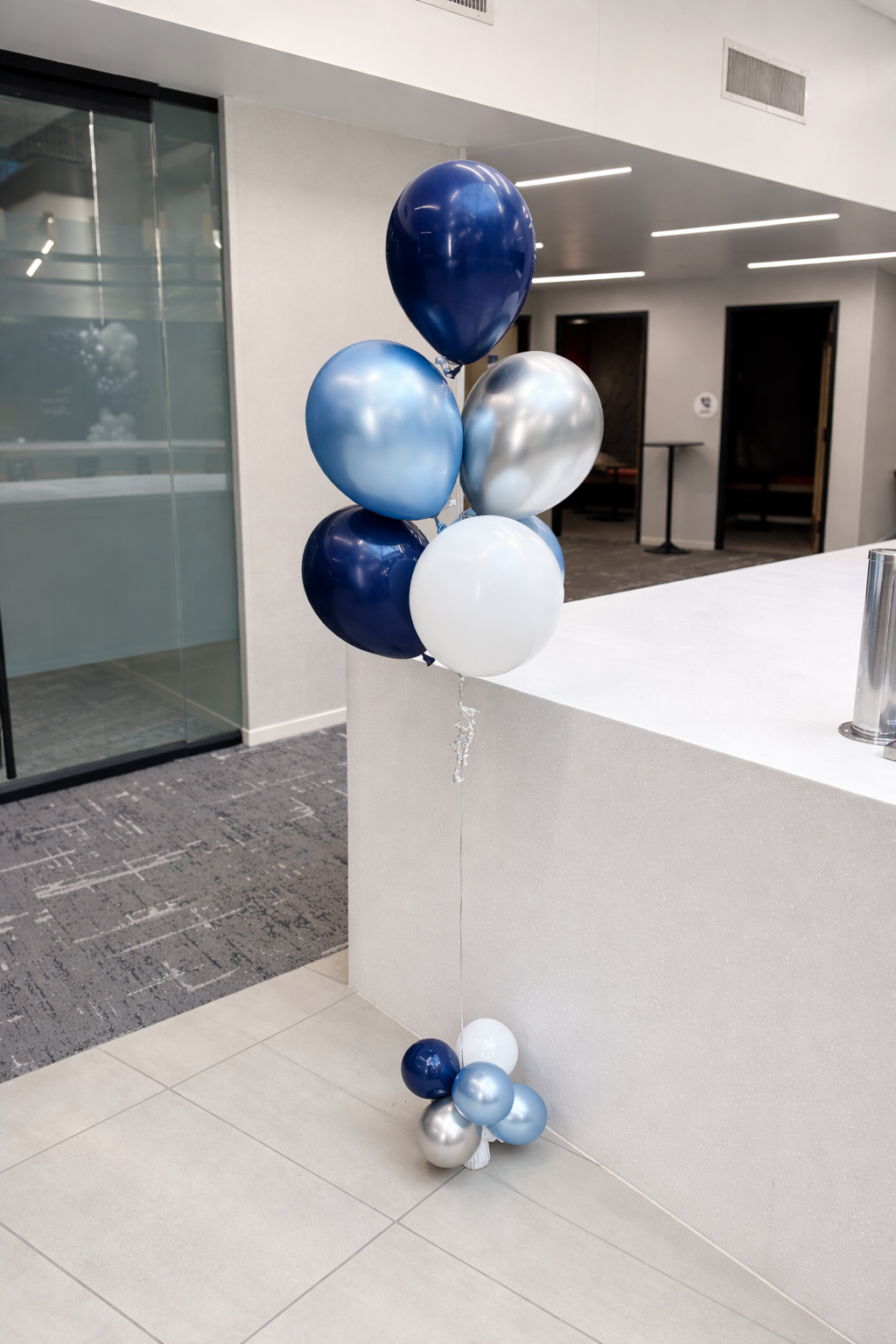 Cluster of blue, white, silver, and navy balloons tied together and placed near a white reception desk in a modern, indoor setting.