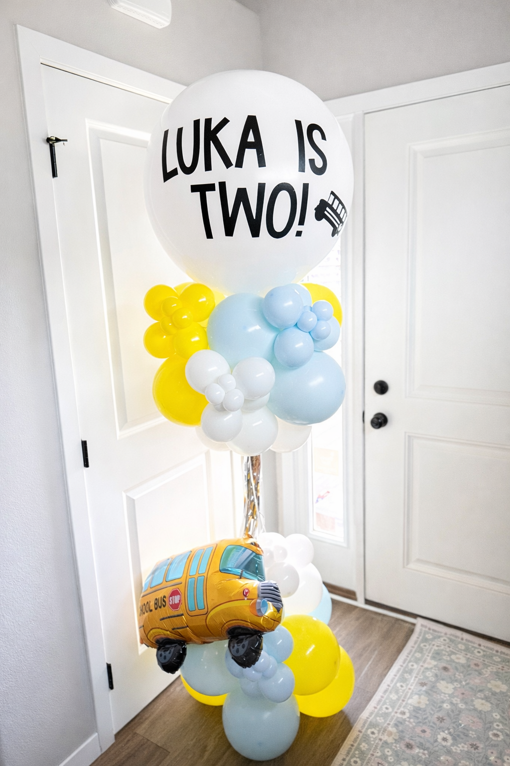 Balloon arrangement at the entrance featuring a large white balloon with 'LUKA IS TWO!' text and a school bus shaped balloon, with yellow, blue, and white balloons.
