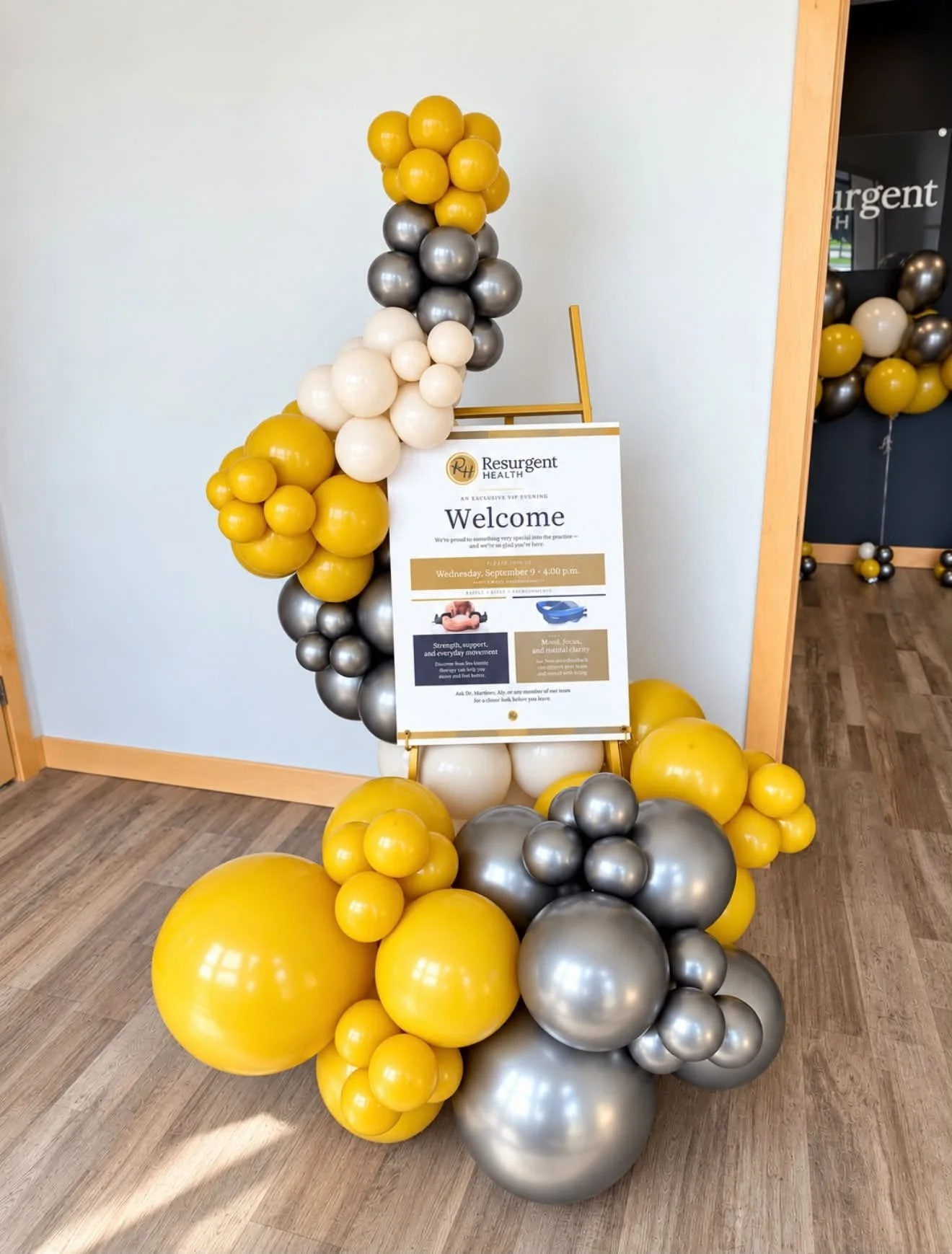 Decorative balloon arrangement with yellow, silver, black, and white balloons surrounding a sign for Resurgent Health.