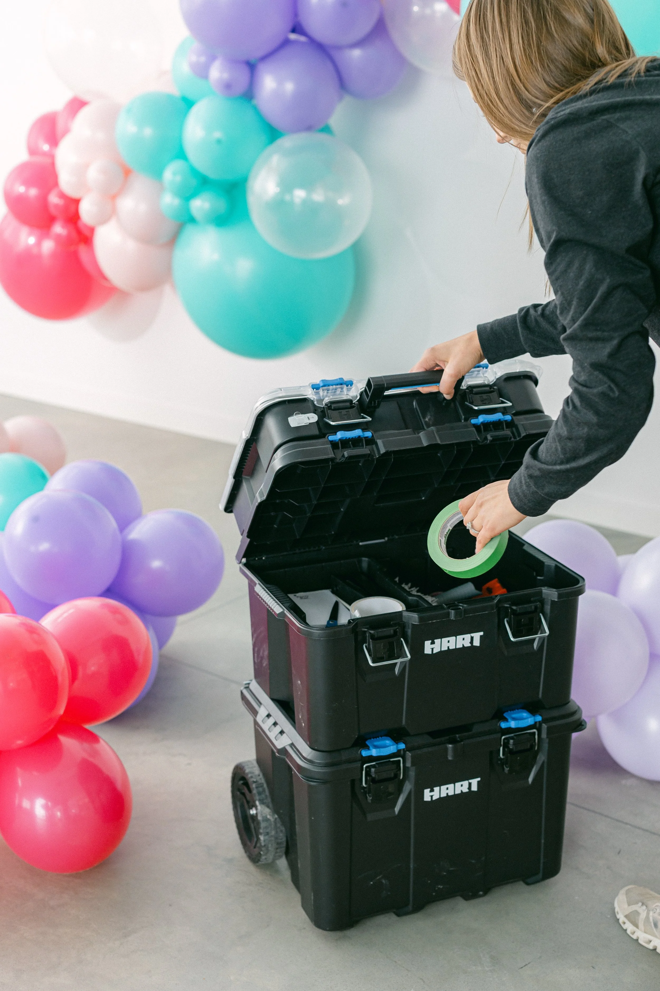 A woman arranging colorful balloons on a white wall, with a black toolbox on wheels labeled 'HART' nearby. The balloons are in purple, teal, white, pink, and red, creating a festive atmosphere.