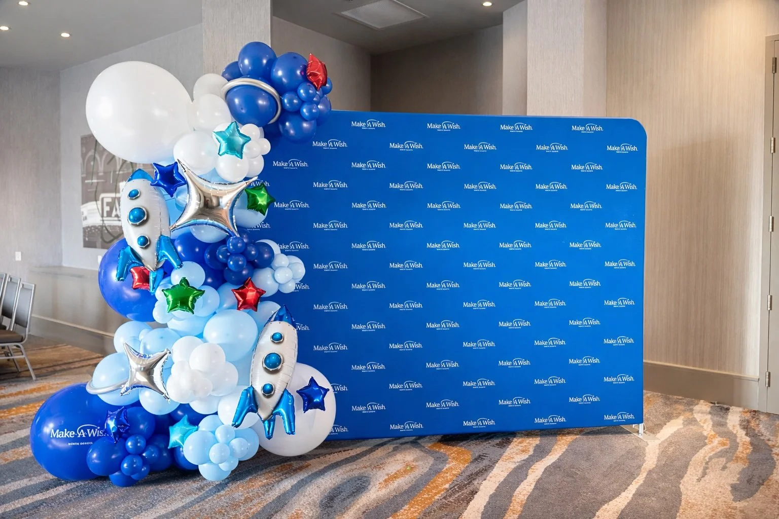 Balloon arrangement with white, blue, red, green, and star-shaped balloons, along with silver rocket-shaped balloons, positioned next to a blue backdrop featuring the 'Make-A-Wish' logo repeated in a pattern.