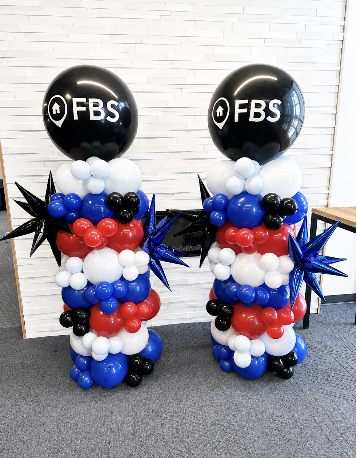 Colorful balloon columns with black, white, red, and blue balloons, topped with large black balloons displaying the FBS logo and icons of a house and location pin, against a white brick wall.