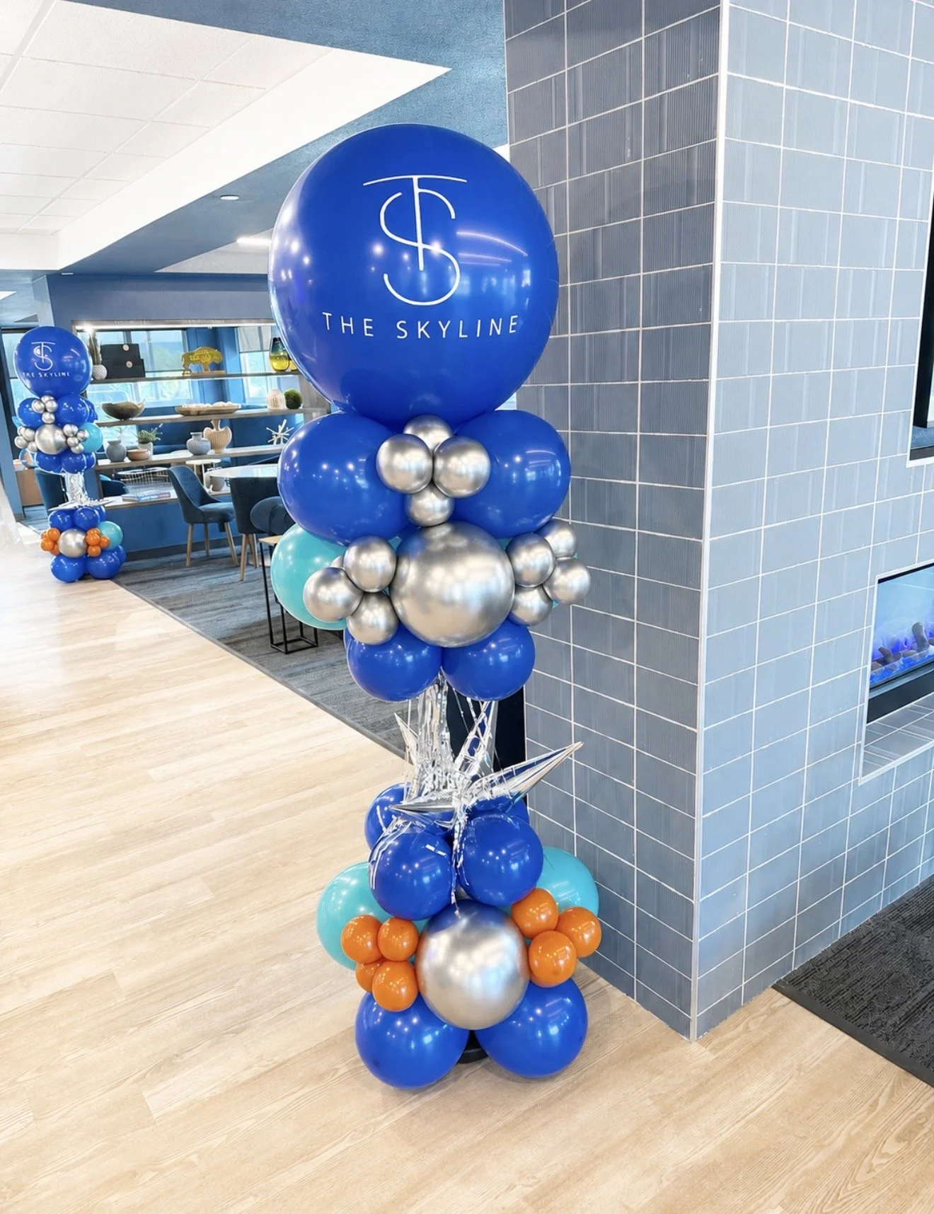 A cluster of balloons in blue, silver, light blue, and orange with a large blue balloon on top reading 'The Skyline' and featuring a stylized logo, displayed in an indoor setting with blue tiles on the wall and tables and chairs in the background.