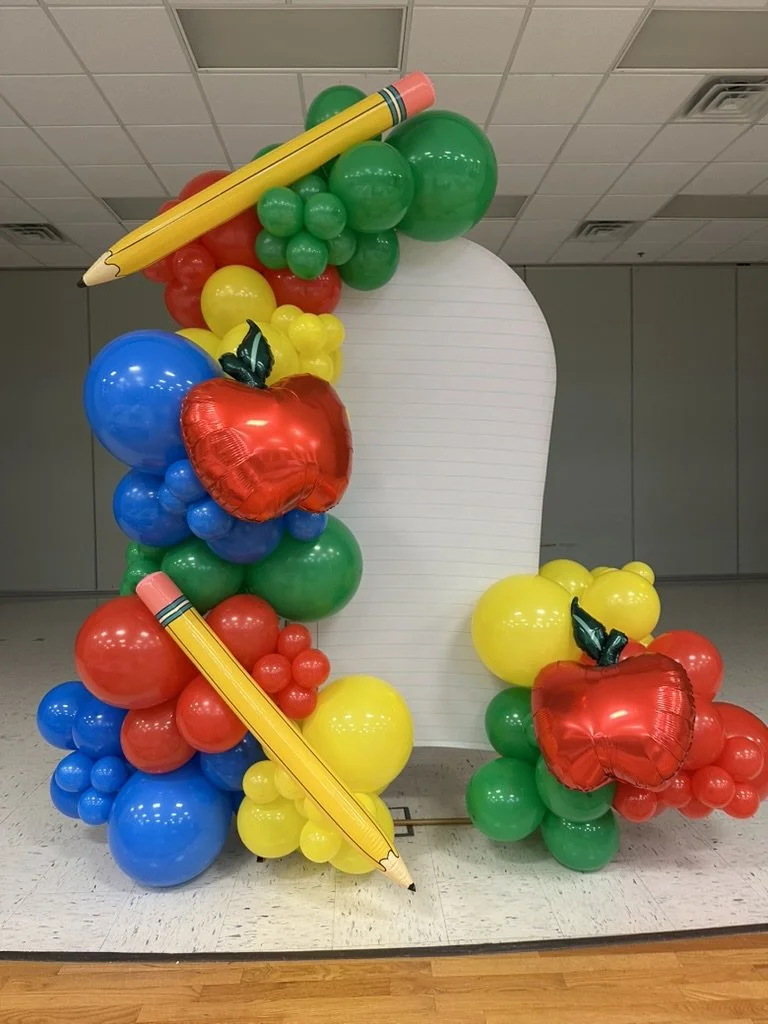 Colorful balloon arch decorated with apple-shaped balloons and large pencil-shaped balloons, set against a plain background.