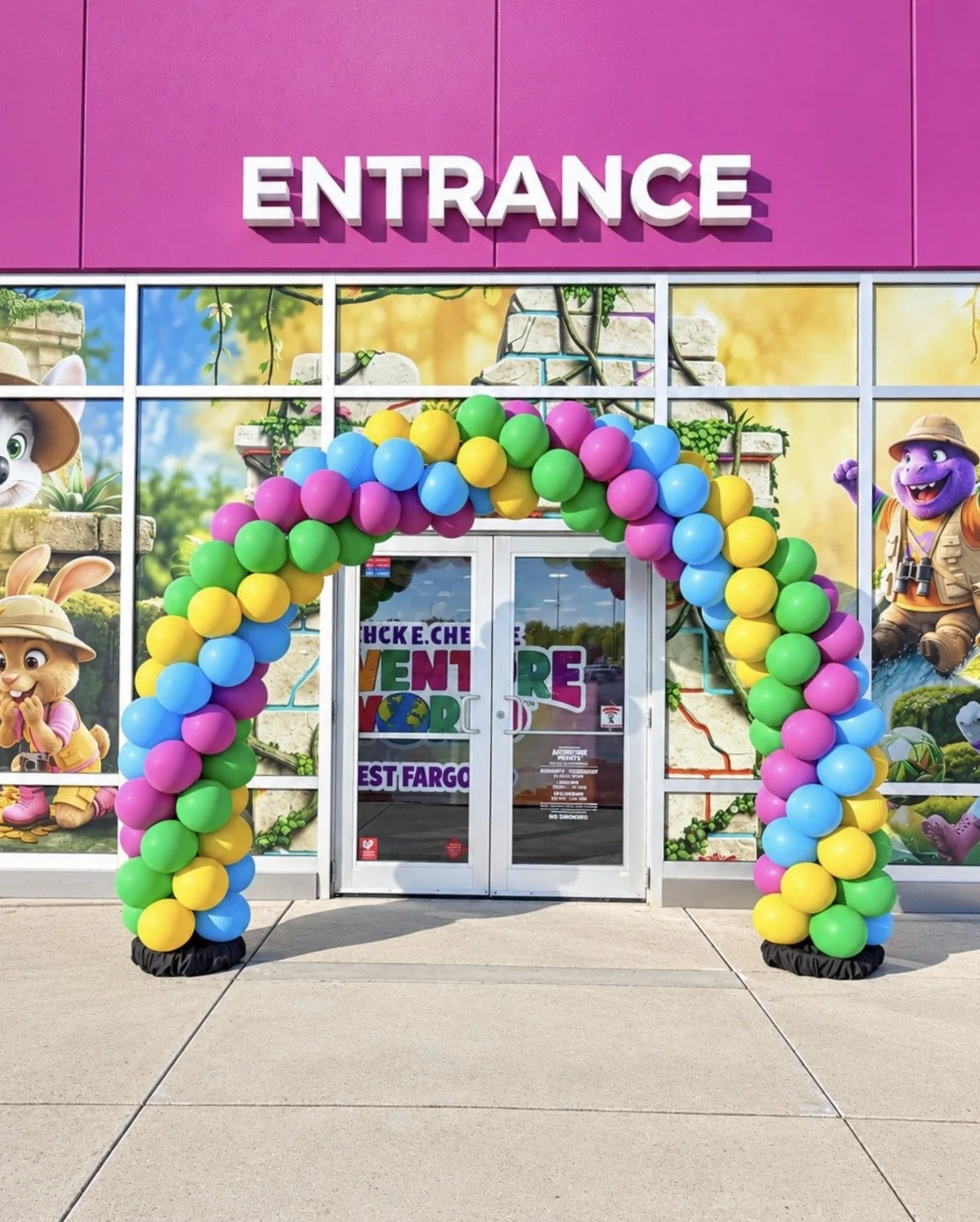 Colorful balloon arch with pink, yellow, green, and blue balloons in front of a store entrance with cartoon animal characters on the window.