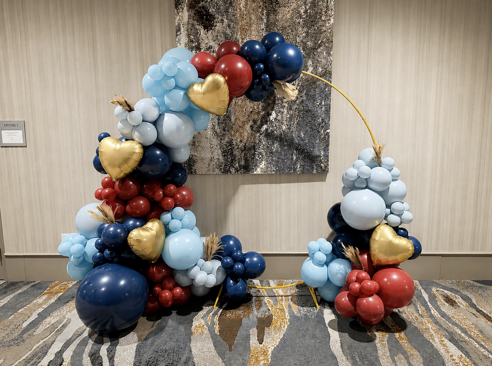 Balloon decoration arch with red, blue, white, and gold balloons, including heart-shaped gold balloons, set against a decorative wall and placed over a patterned carpet.
