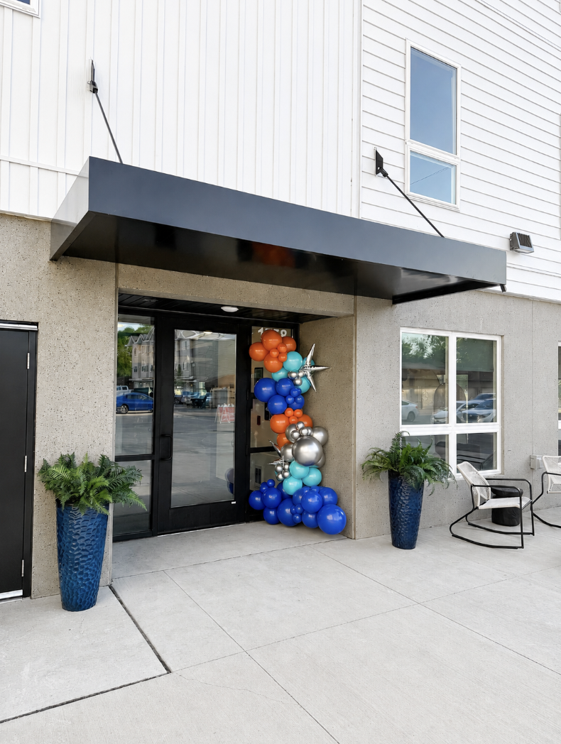 Entrance of a building decorated with balloons in orange, blue, silver, and teal, with two large potted plants flanking the door, and a bench and window on the side.
