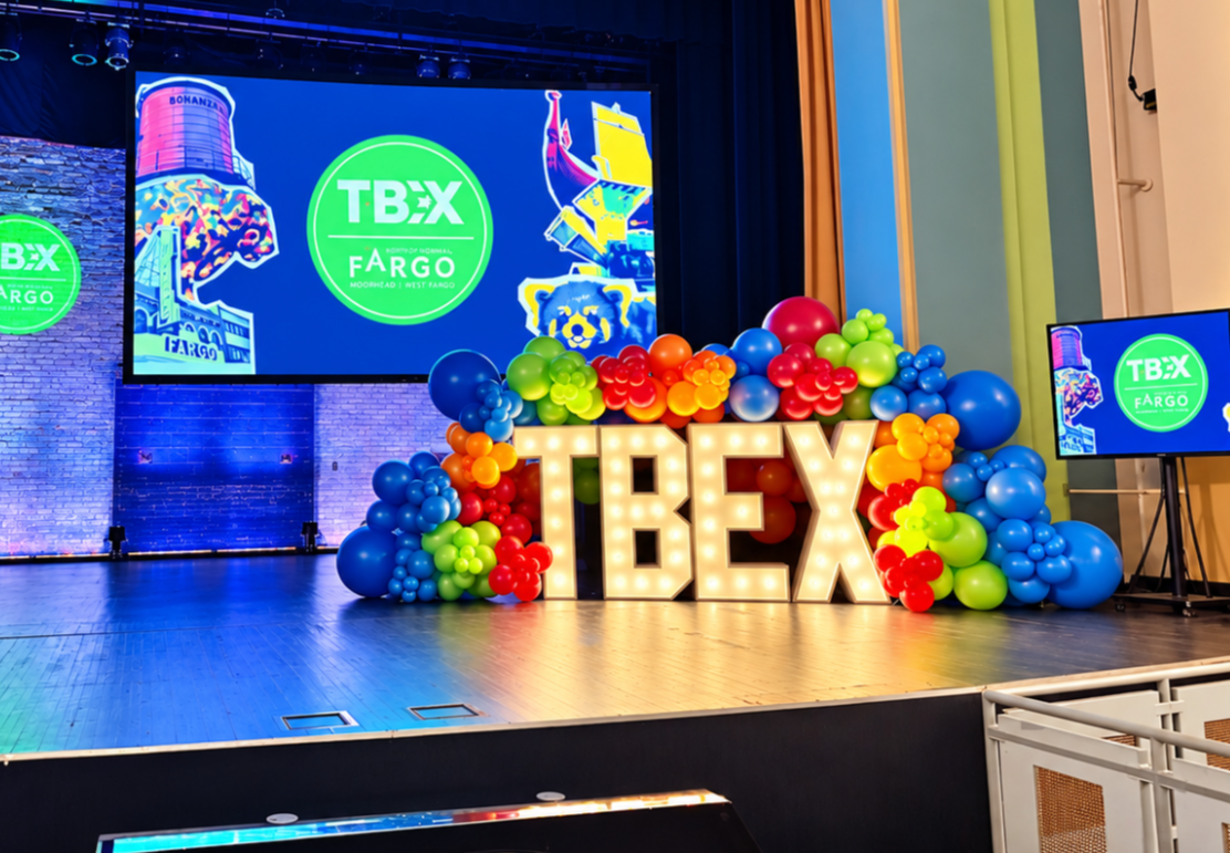 Stage decorated with large illuminated letters spelling T-BEX, surrounded by colorful balloons, large screens displaying the TBEX logo and graphics of Fargo, with a brick wall backdrop.