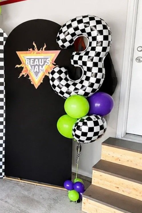 A decorative balloon arrangement featuring a large checkered number three, two green balloons, a purple balloon, and checkered patterned balloons, positioned beside a black backdrop with a "Beau's Jam" logo, near wooden stairs.