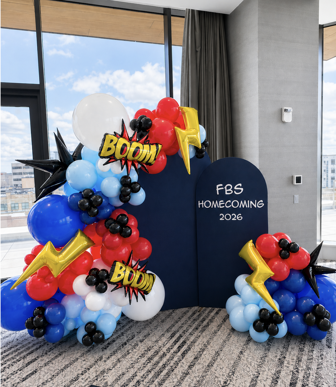 Decorative balloon arrangement with comic book style "BOOM!" signs, lightning bolt balloons, and star-shaped balloons, set up for FBS Homecoming 2026 event indoors near large windows.