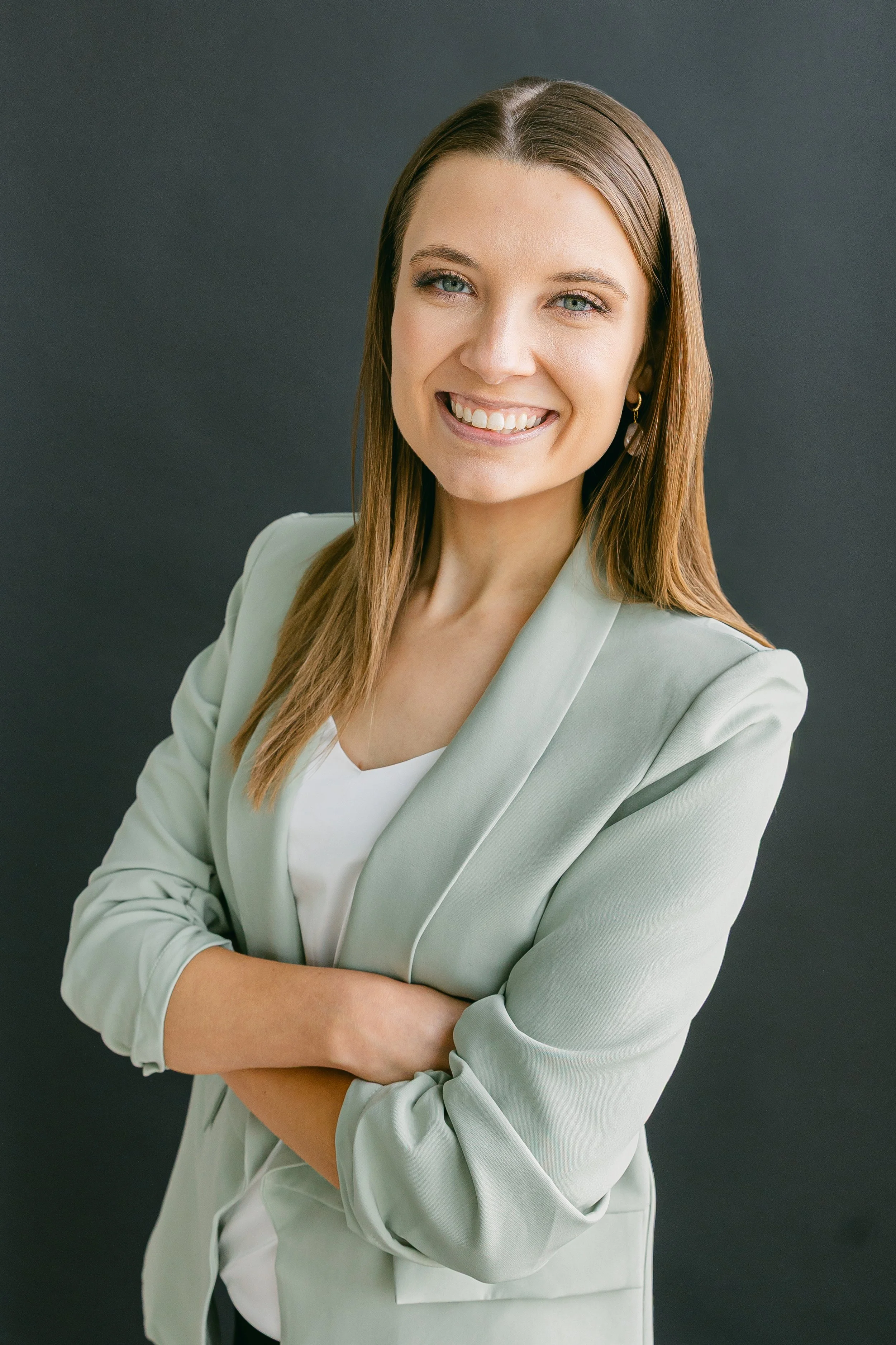 A young woman with light brown hair, smiling, wearing a light grey blazer over a white top, arms crossed, standing against a dark background.