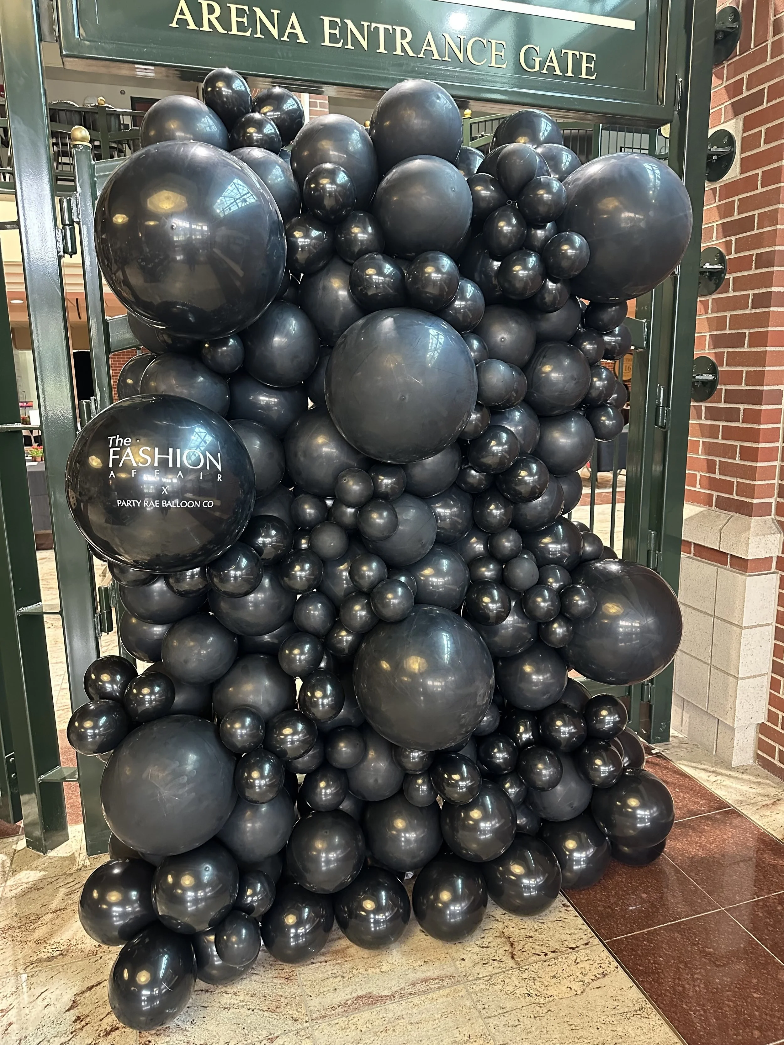 A large decorative balloon display with black balloons of various sizes, situated in front of an arena entrance gate. A black balloon in the display has white text reading "The Fashion Affair x Party Rae Balloon Co."