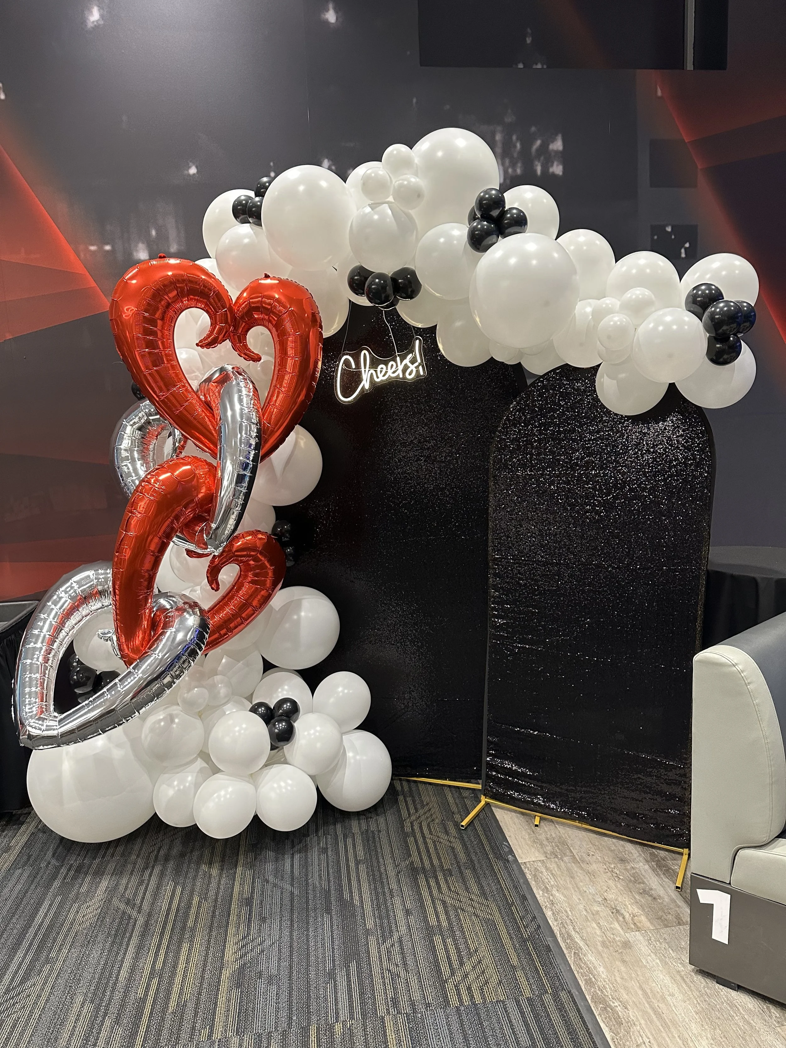 Decorative balloon arrangement with red, silver, black, and white balloons, including a heart-shaped balloon, a neon sign saying 'Cheers!', and a black glittery backdrop. King Pinz Social Fargo