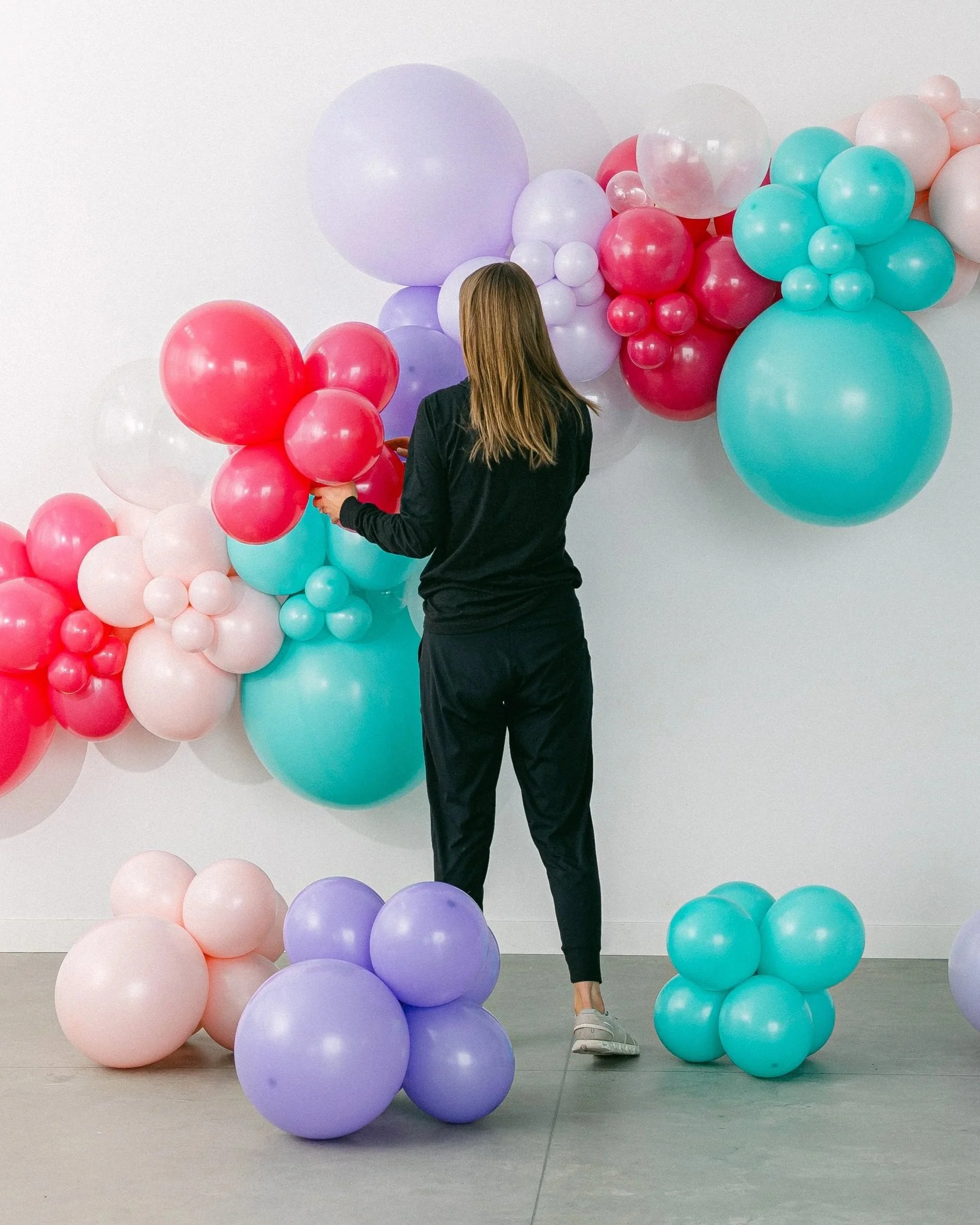 A person with long hair, dressed in dark clothing and sneakers, stands in front of a colorful balloon art installation on a plain white wall.