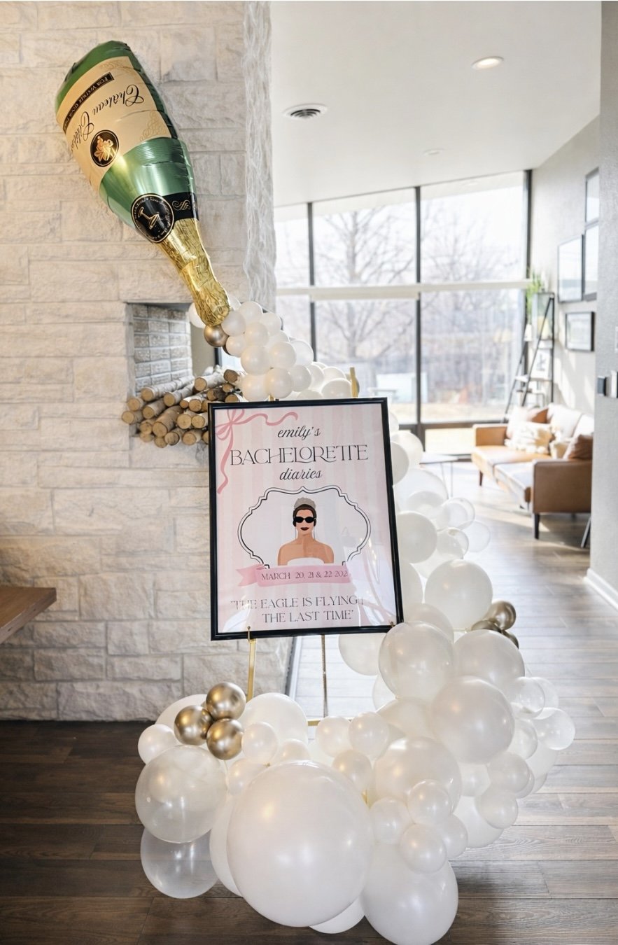 Decorative display for Emily's bachelorette diaries, featuring a large champagne bottle-shaped balloon, white and metallic gold balloons, and a poster with a woman in sunglasses, dates March 20-22, 2022, and a quote about an eagle.
