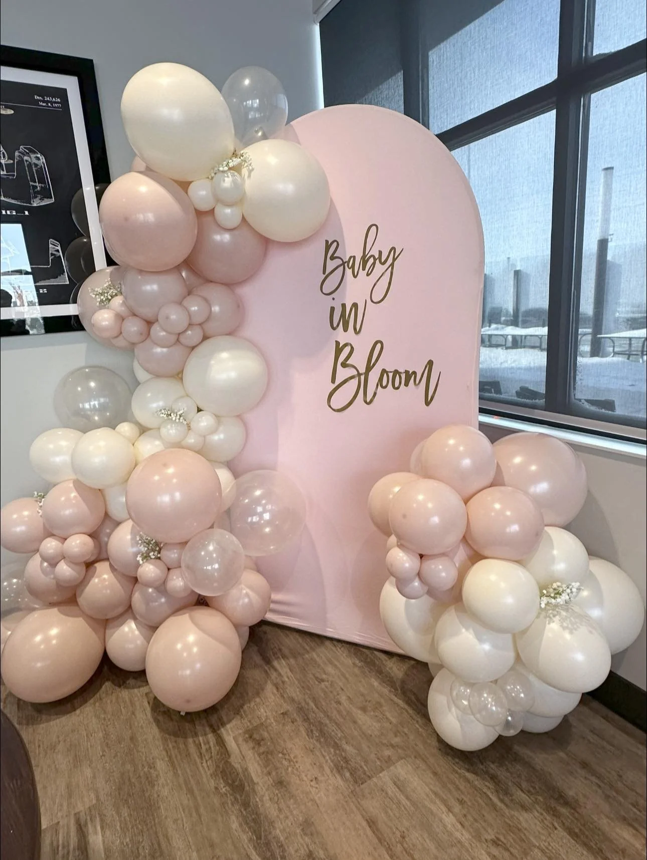 Decorative balloon arrangement with pink, white, and clear balloons surrounding a pink backdrop that reads 'Baby in Bloom' in gold lettering, set indoors near a window.