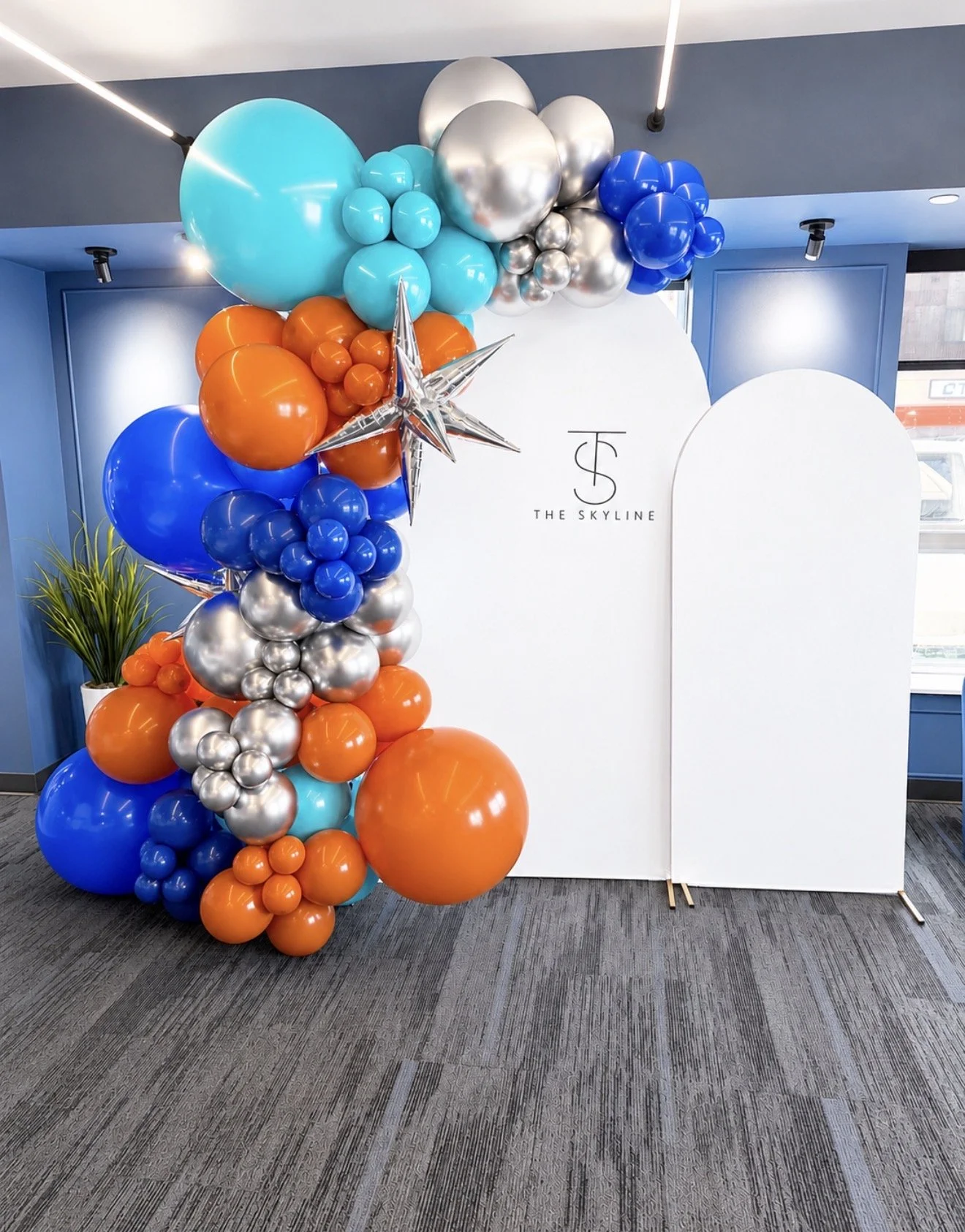 Decorative balloon arrangement with orange, blue, silver, and turquoise balloons, along with star-shaped metallic balloons, standing next to a white partial wall with a logo that reads 'The Skyline' in black text.