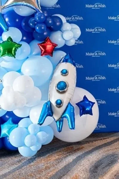 Rocket-shaped balloon surrounded by blue, white, green, and red star-shaped and round balloons, with a blue Make-A-Wish backdrop.