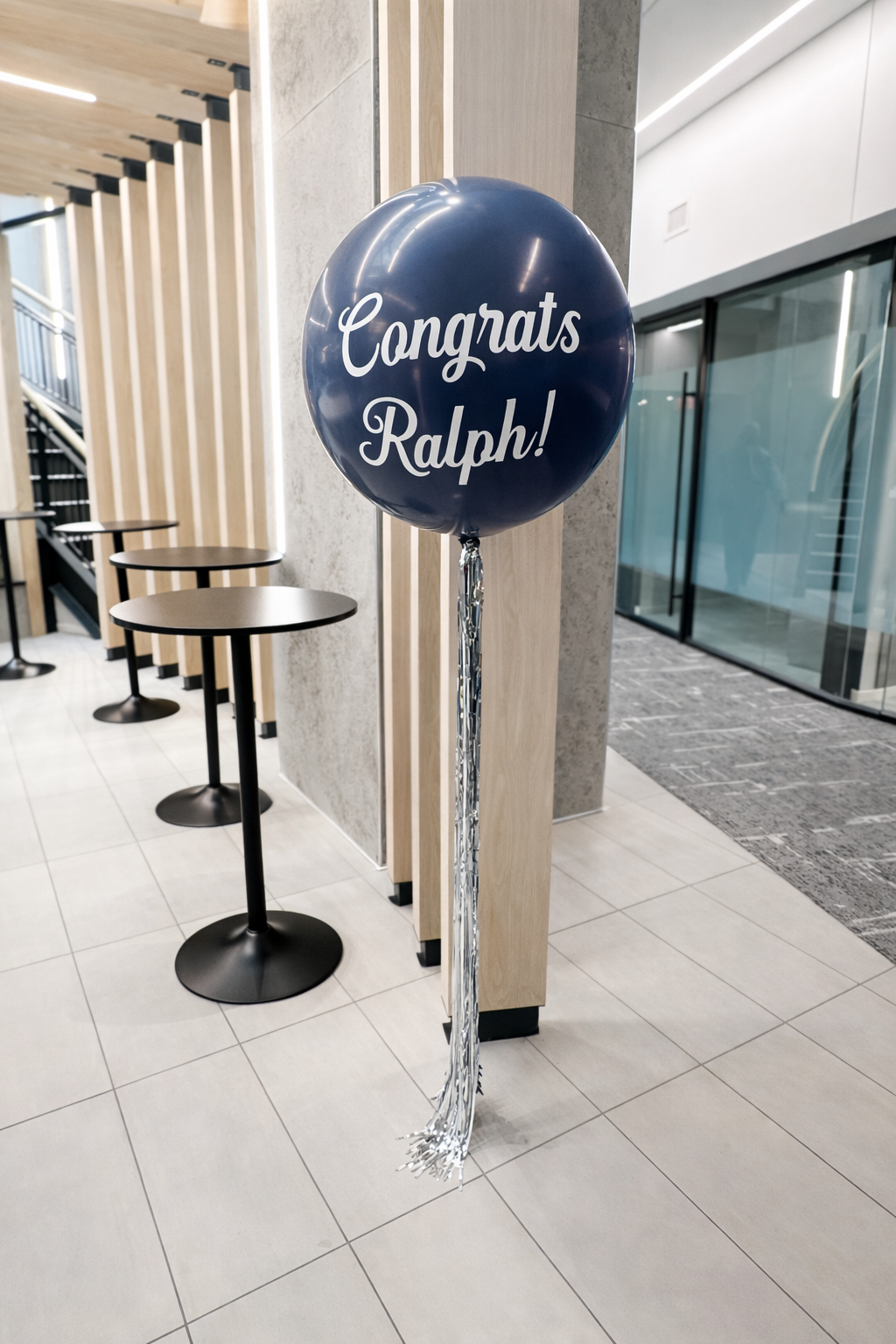 Blue balloon with white text saying "Congrats Ralph!" tied to a silver weight, standing in a modern indoor space with tables and a wooden ceiling.