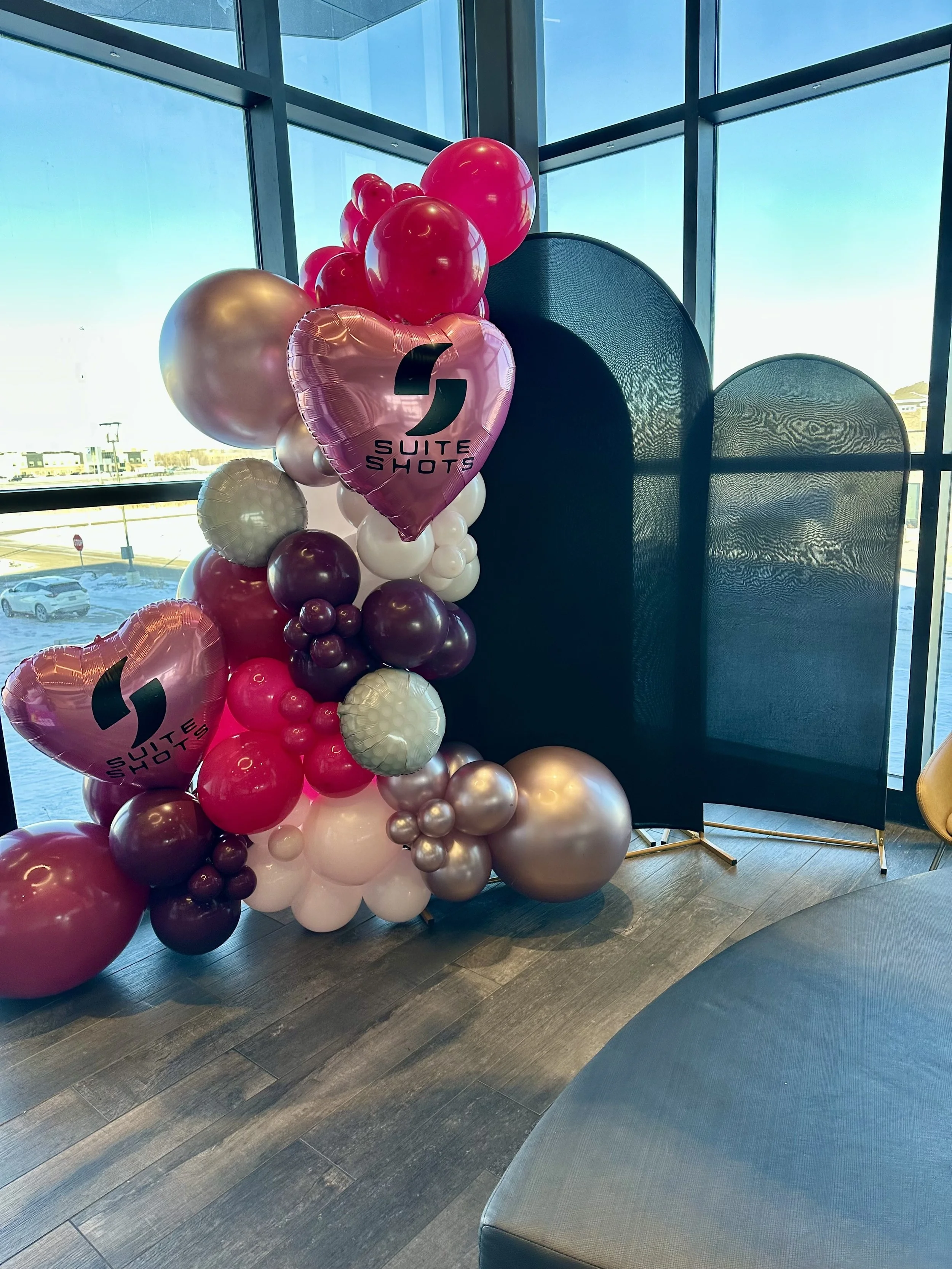 Decorative balloon arrangement with pink, white, gold, and maroon balloons, including heart-shaped balloons with the Suite Shots logo, placed near a window in a modern indoor setting.