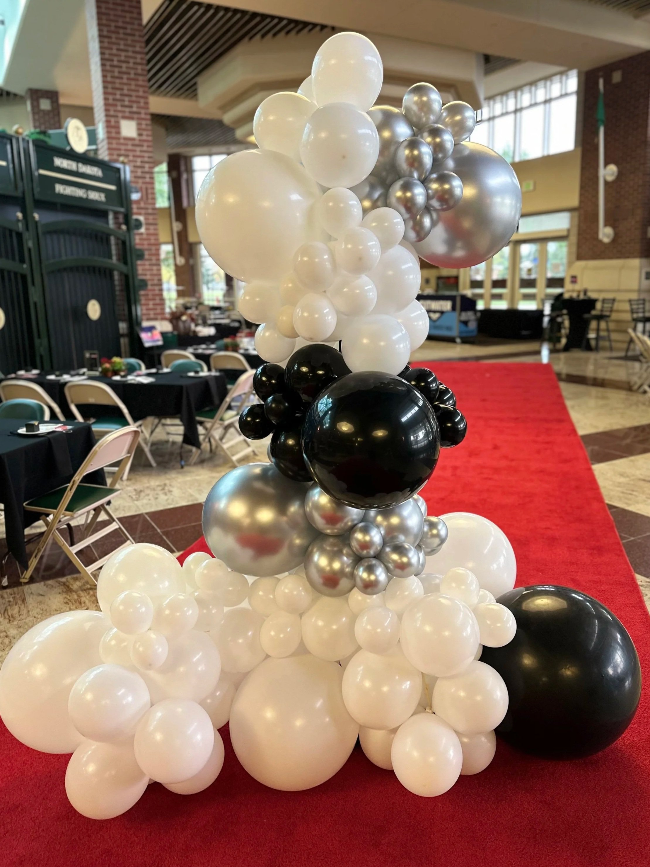 Decorative balloon arrangement with black, white, and silver balloons on a red carpet in a banquet hall.