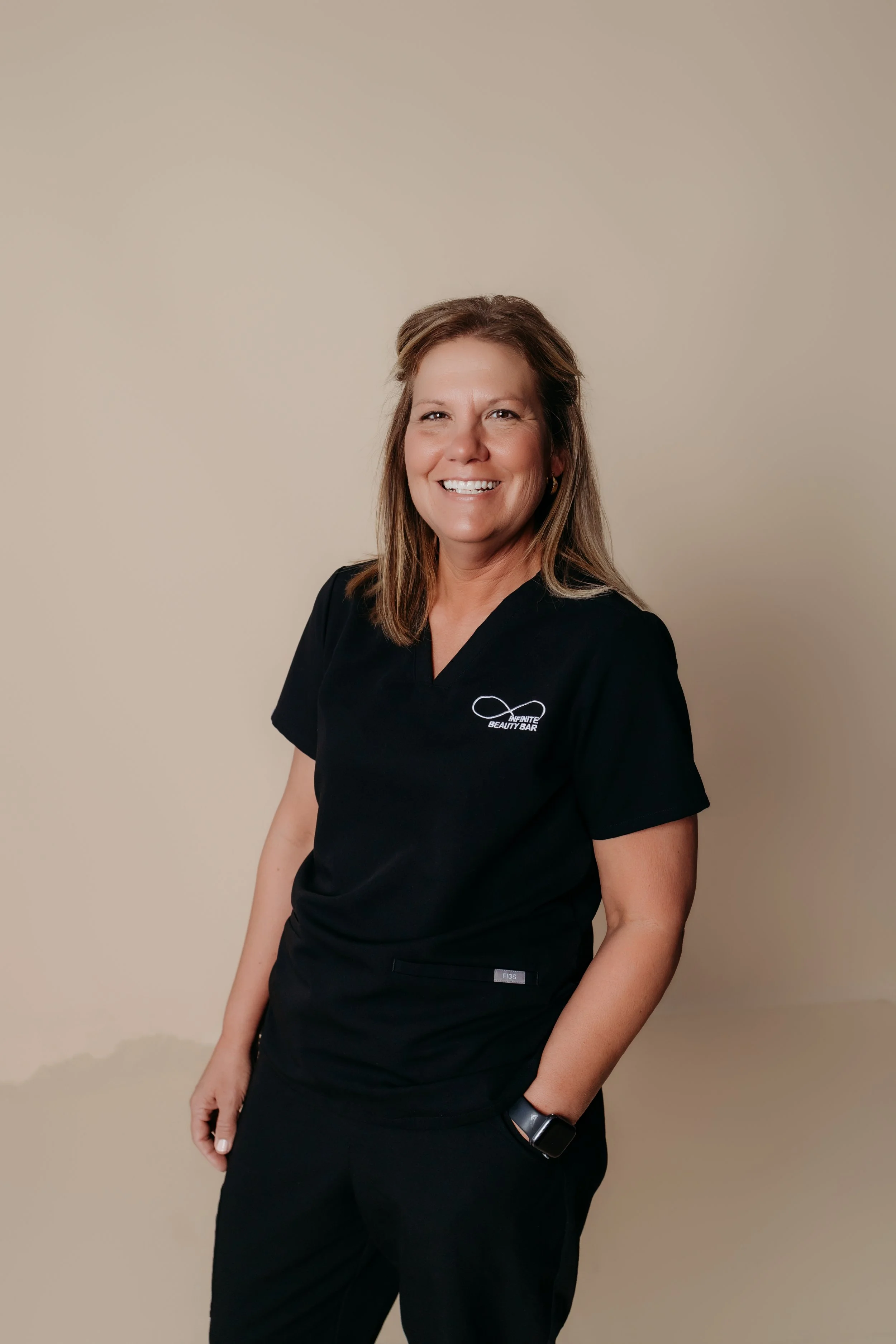 Alissa, RN at Infinite Beauty Bar, has long, wavy brown hair, green eyes, and is wearing a black top and a necklace with a pendant.