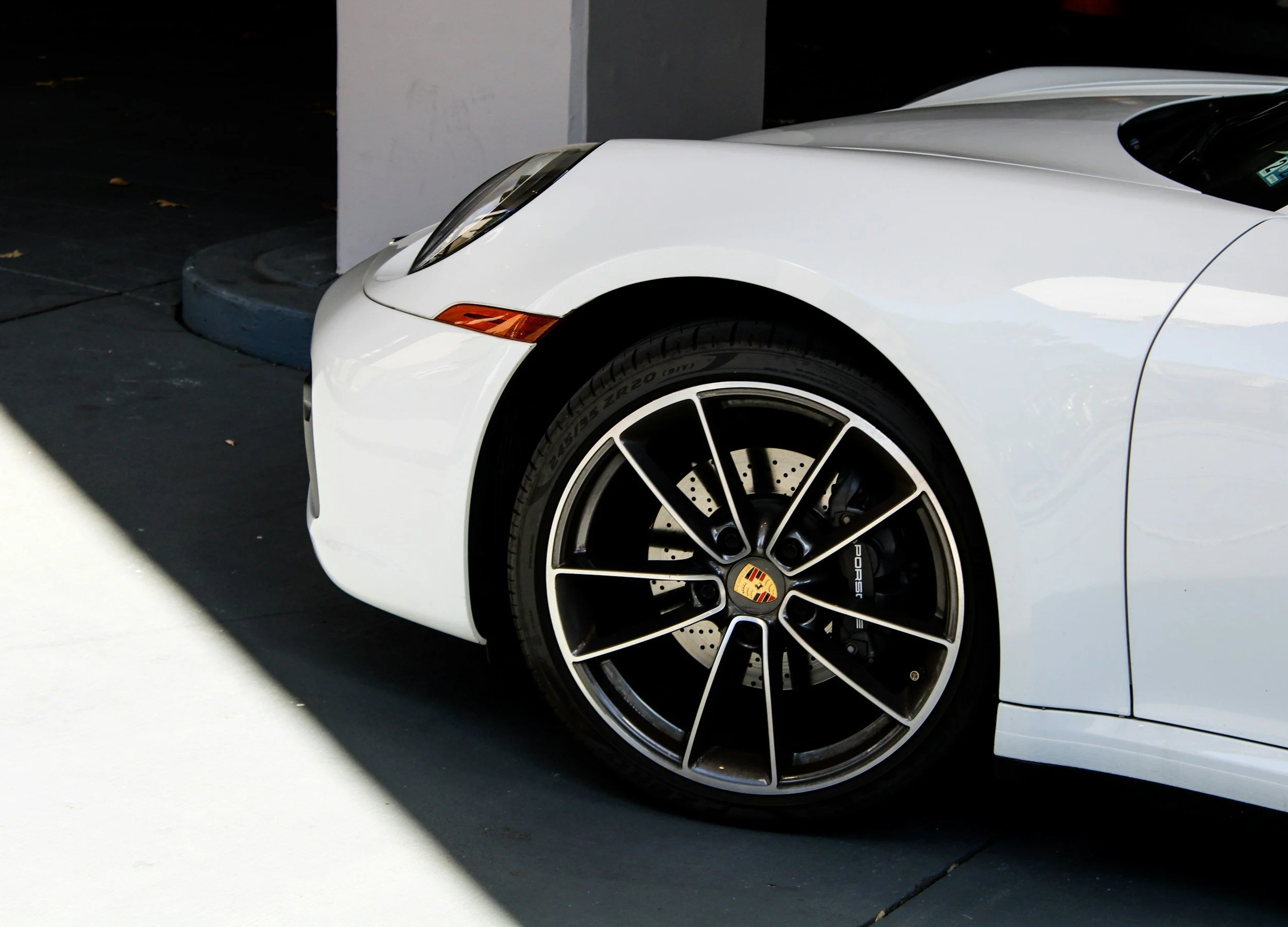 Close-up of the front part of a white Porsche sports car, focusing on the wheel and tire, parked near a light gray wall and a concrete curb.