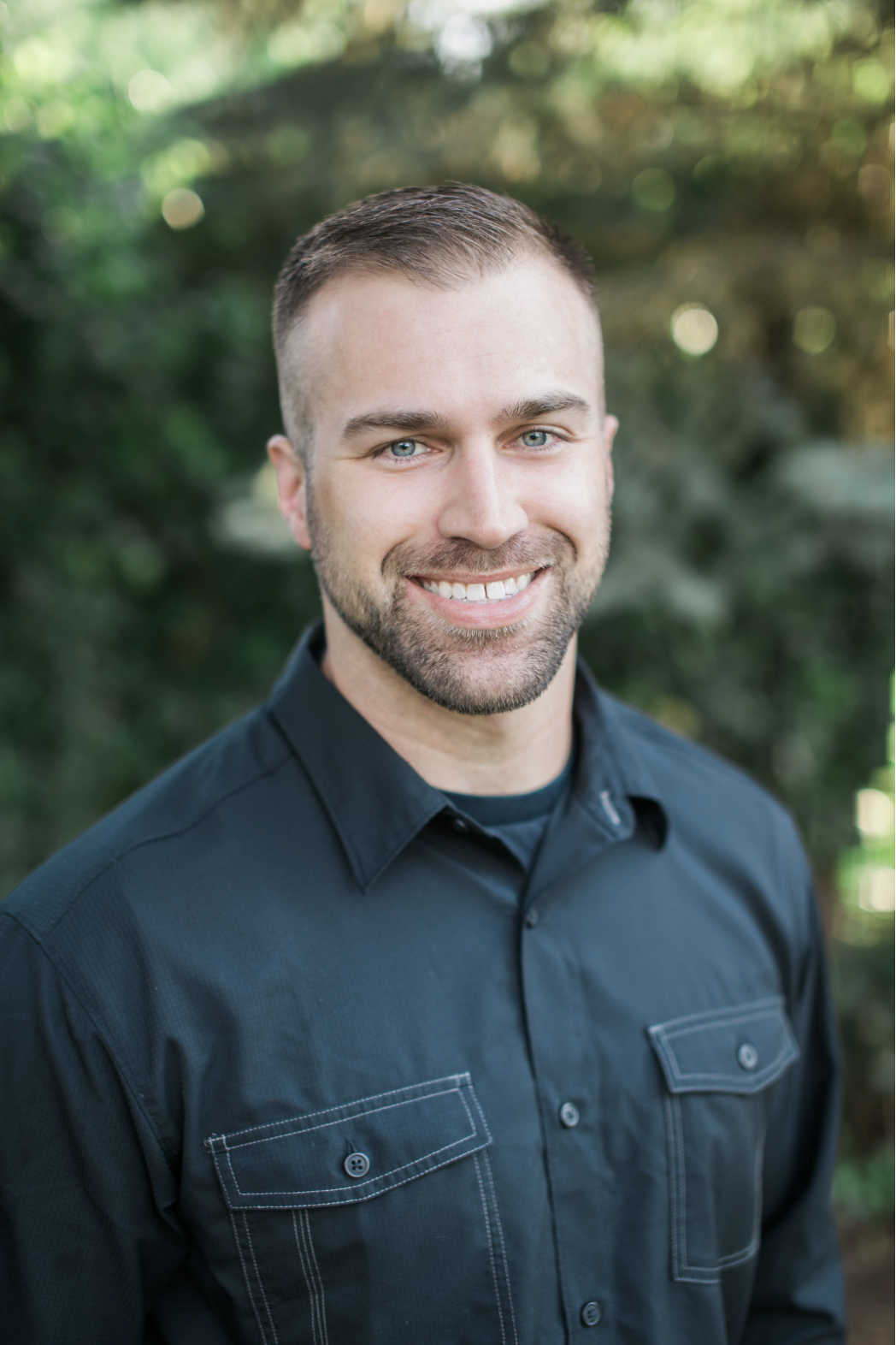 Headshot of a smiling man with short brown hair and a beard, wearing a dark button-up shirt, outdoors with a blurred green background in Medford, Oregon.