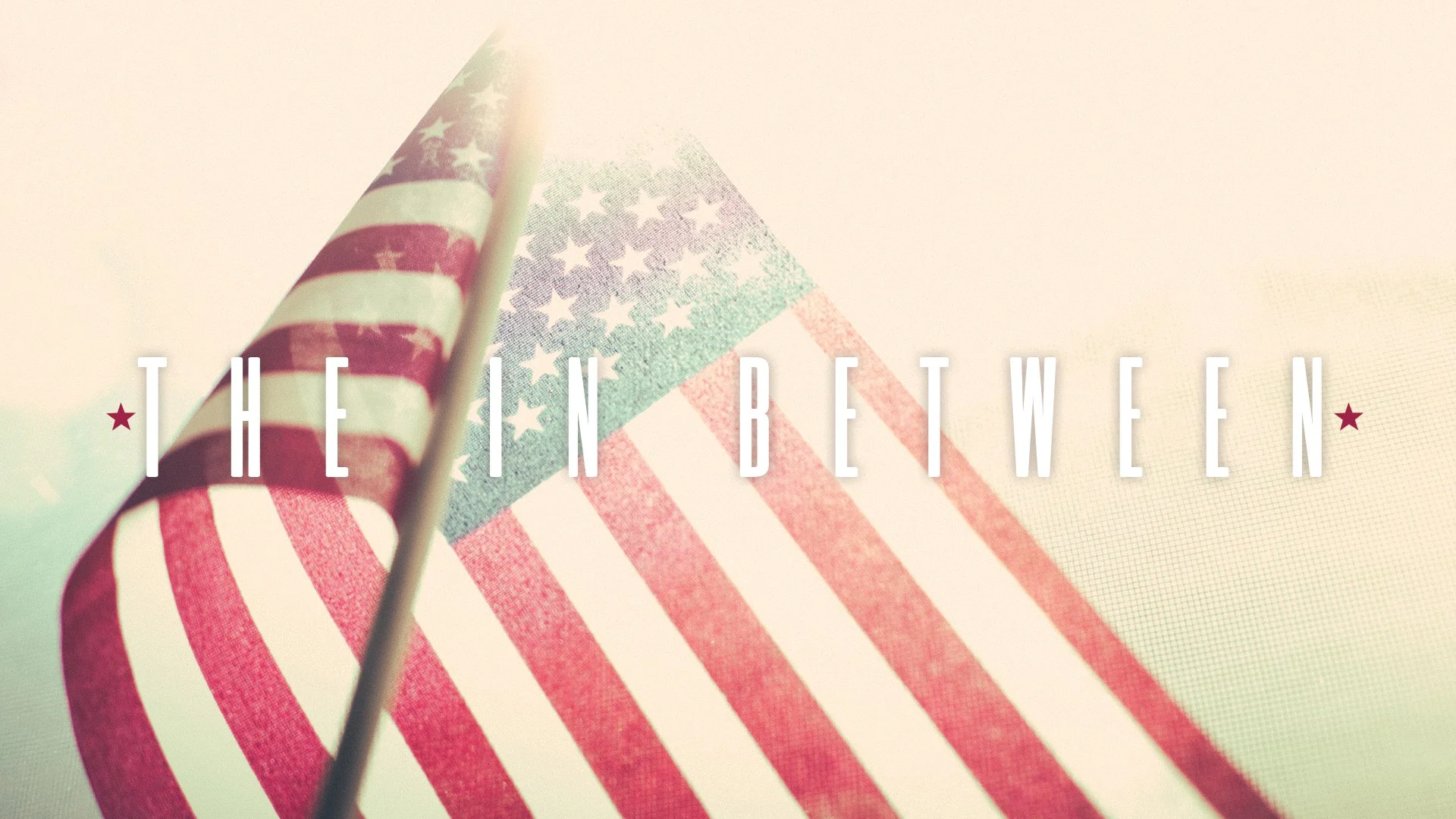 The image shows a paper American flag with red and white stripes and a blue field with white stars, set against a light background. The words "THE IN BETWEEN" are overlaid in white letters, with red stars on either side of the text.