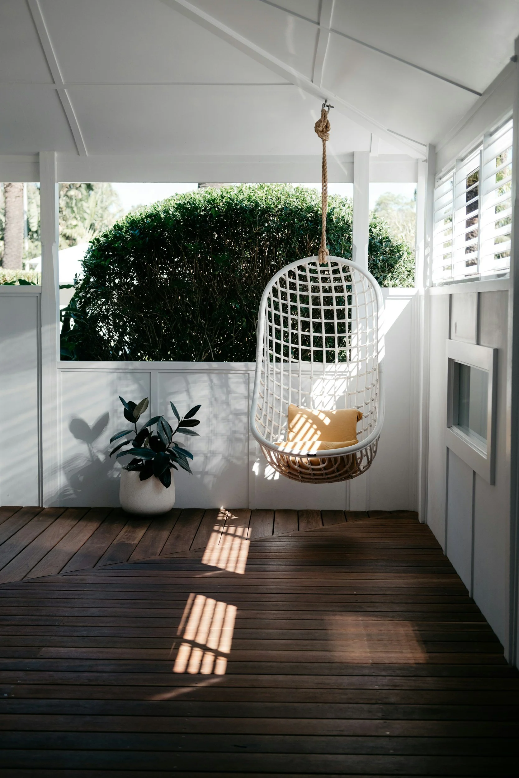 A wooden porch with white walls and a hanging white wicker chair with a yellow cushion. A large green shrub is in the background, and sunlight casts shadows through window blinds onto the floor, with a potted plant in a white ceramic pot nearby.