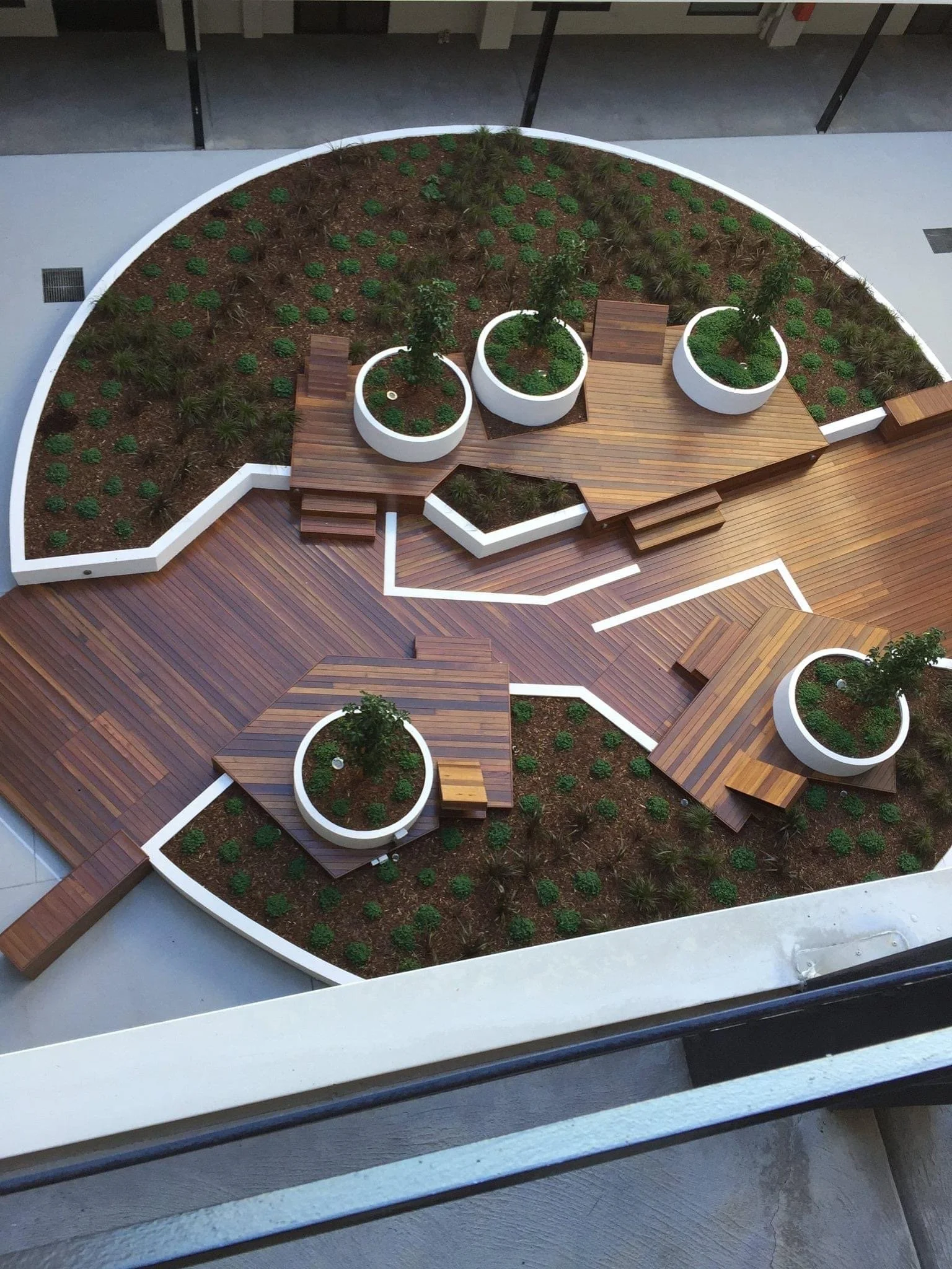 Bird's eye view of a modern rooftop garden with wooden decking, circular planters with trees, and various small plants.