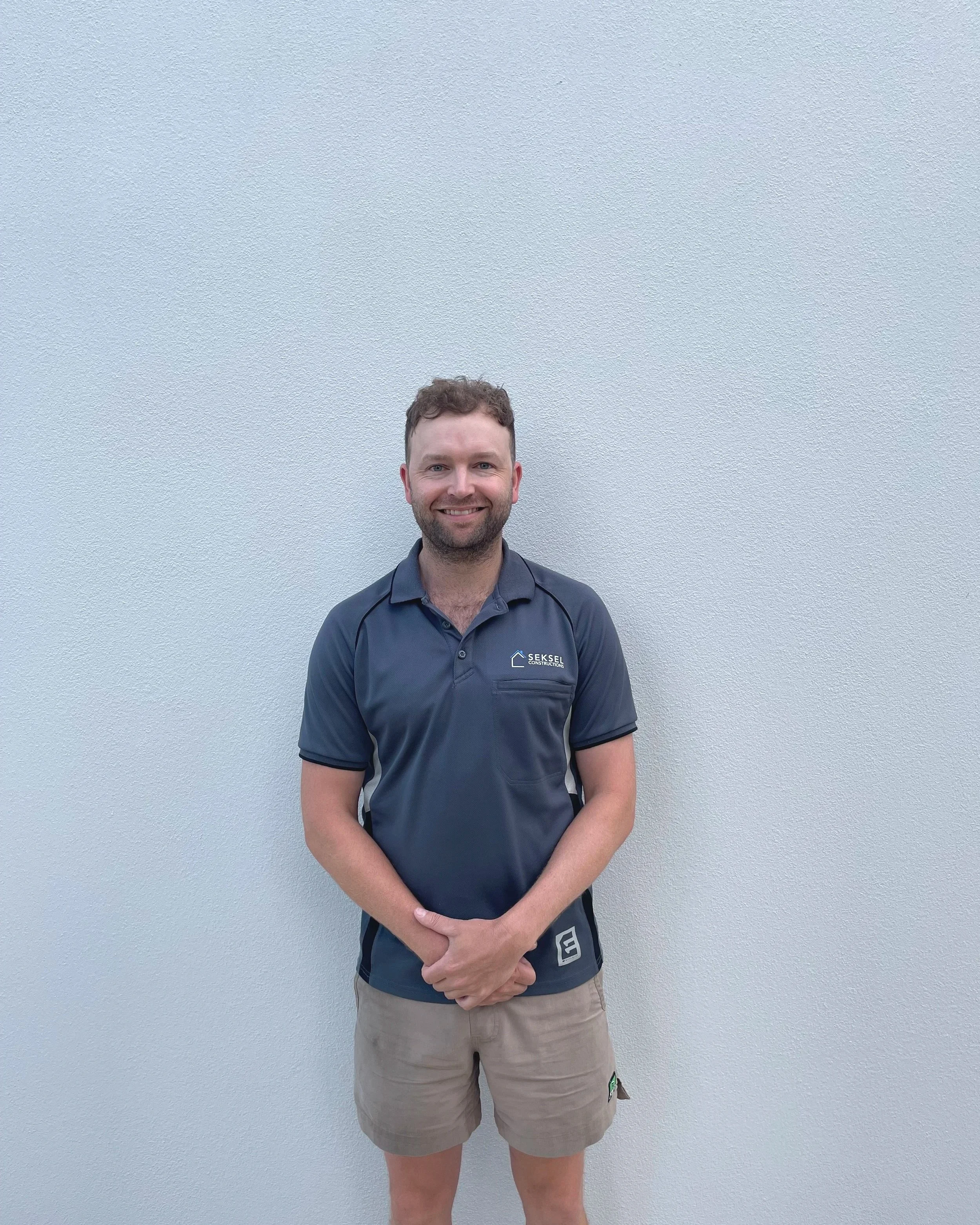 A man with a beard and curly hair smiling, wearing a navy blue polo shirt with a logo on it and beige shorts, standing in front of a plain white wall.