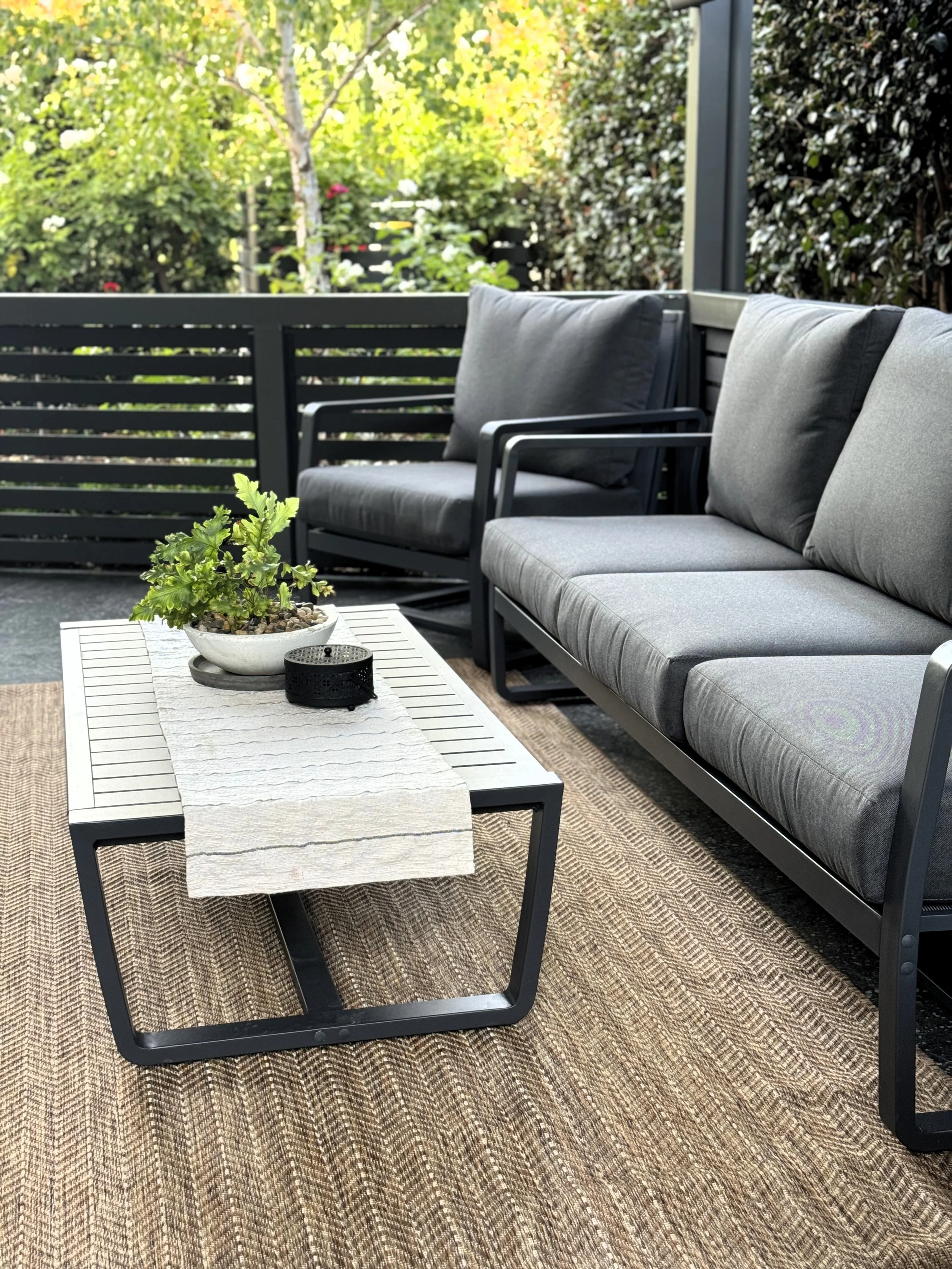 Outdoor patio with gray cushioned seating, a white slatted coffee table with potted plant and black decorative container, beige rug, surrounded by greenery and a black wooden fence.