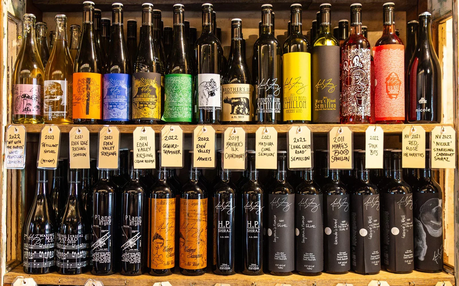 A wooden shelf displaying multiple bottles of wine with handwritten tags indicating their types and vintages. The bottles vary in color and label design, including reds, whites, and rosés, with tags showing details like '2022 Off with the Fairies', '
