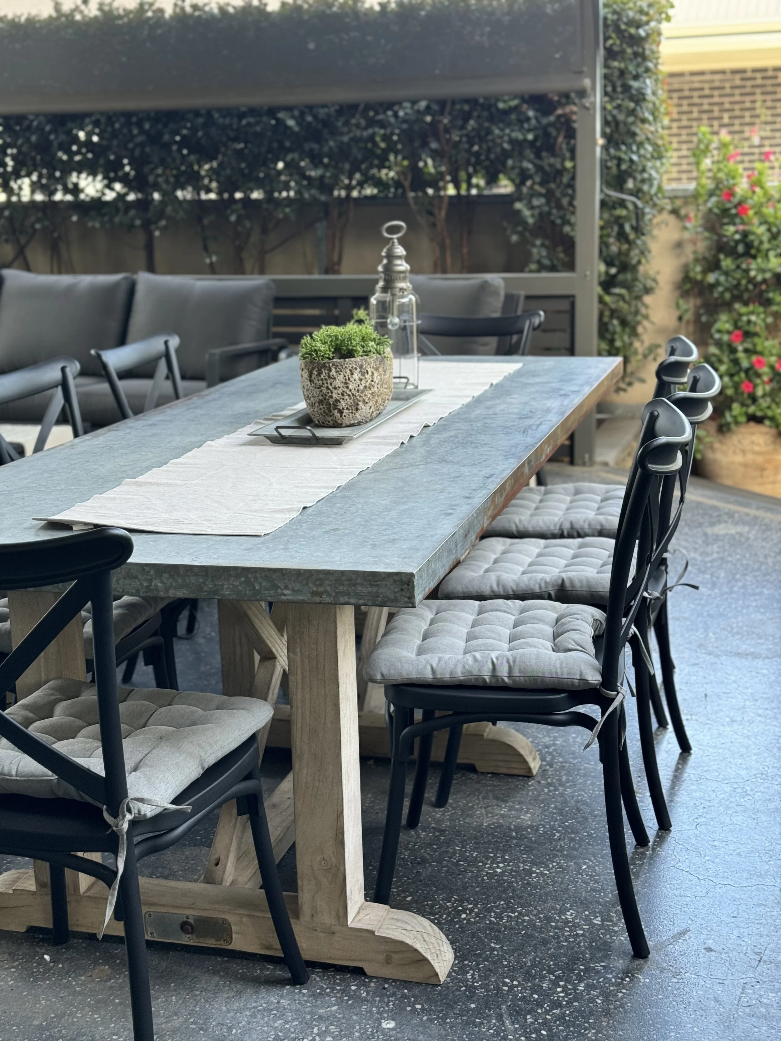 Outdoor dining table with gray and beige chairs, a white table runner, a potted plant, and decorative lanterns, surrounded by green plants and flowers.