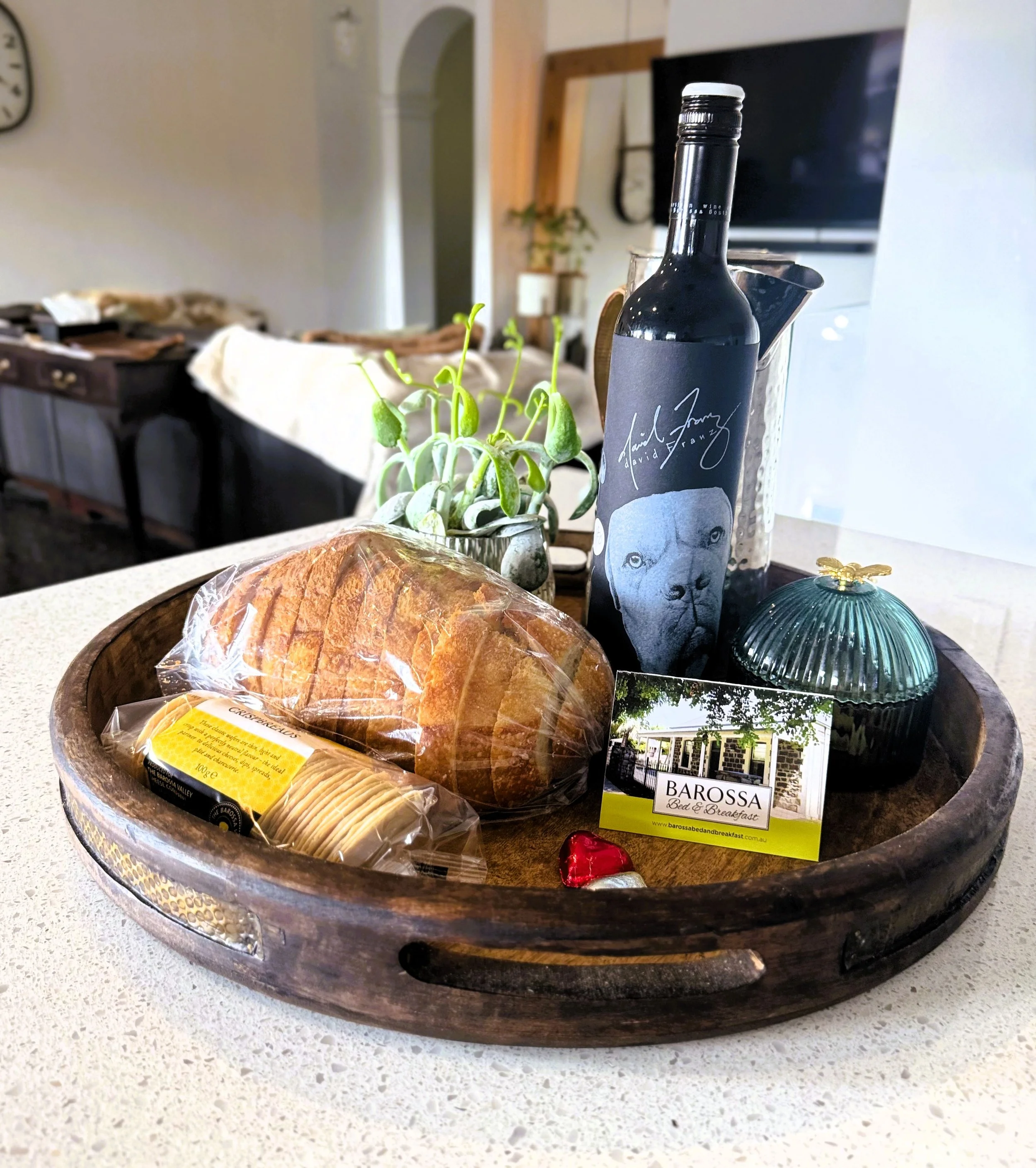 A round wooden tray on a white kitchen countertop holding a loaf of bread in plastic, cheese, a bottle of wine with a black label, a small potted plant, a decorative box with a bee on top, a business card for Barossa Bed & Breakfast, and some wrapped