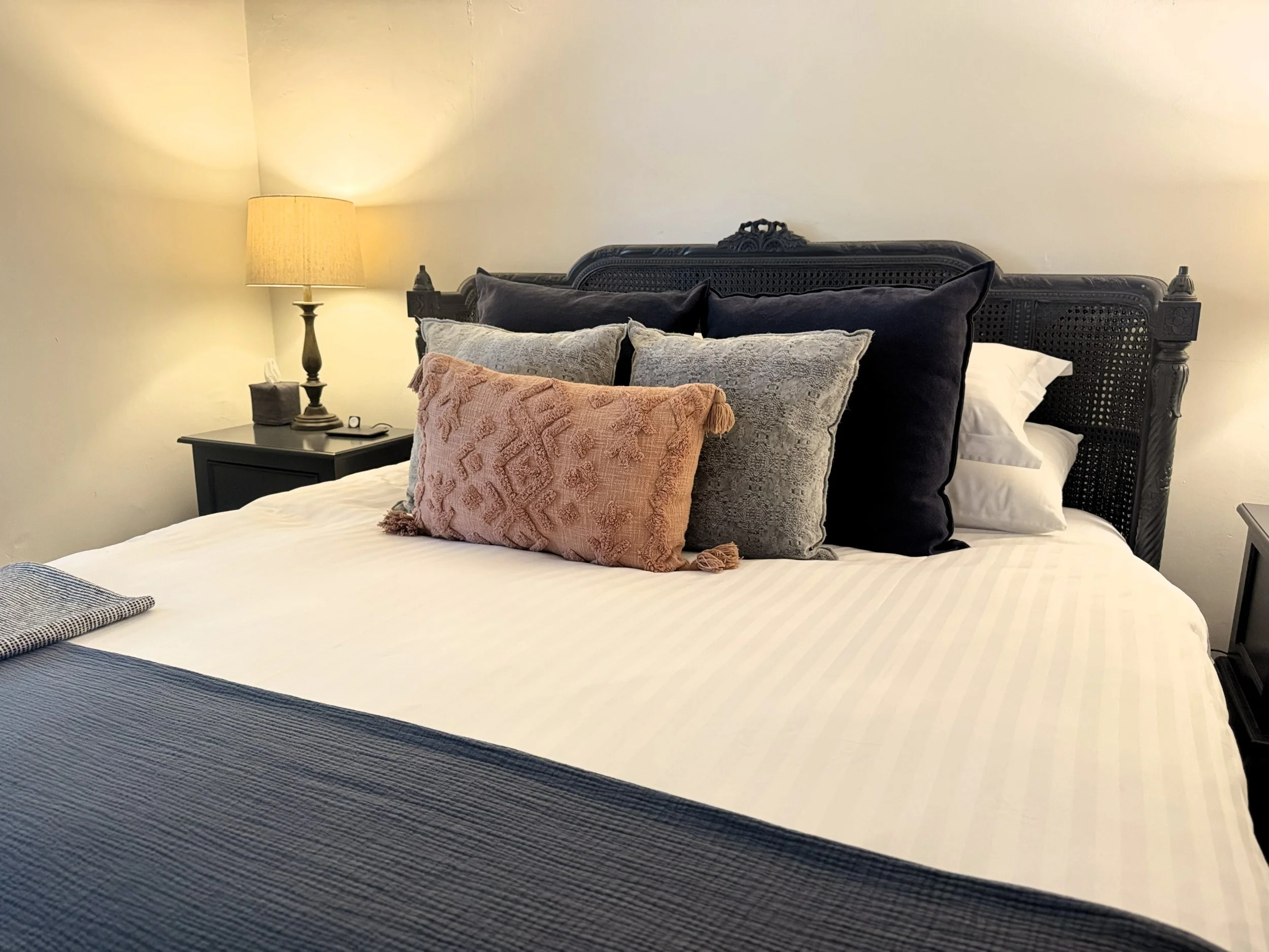 A neatly made bed with decorative pillows, black headboard, bedside table with lamp, tissue box, and a phone at night.